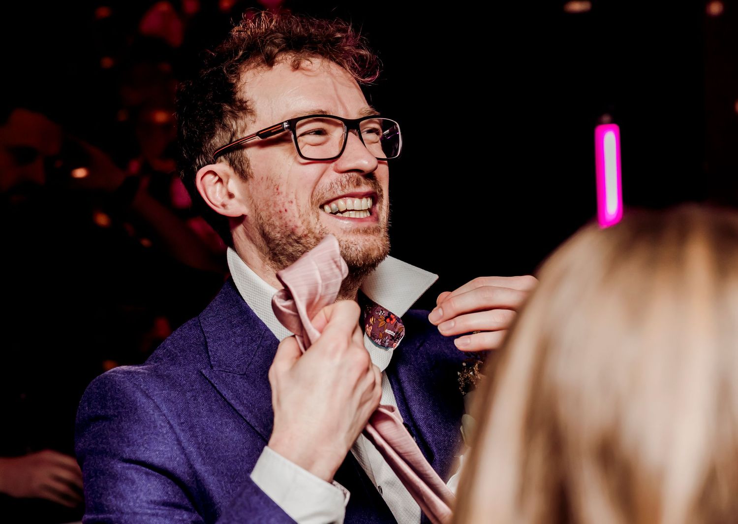 Smiling groom in a blue suit adjusts his tie at a wedding reception with pink neon lights in background.