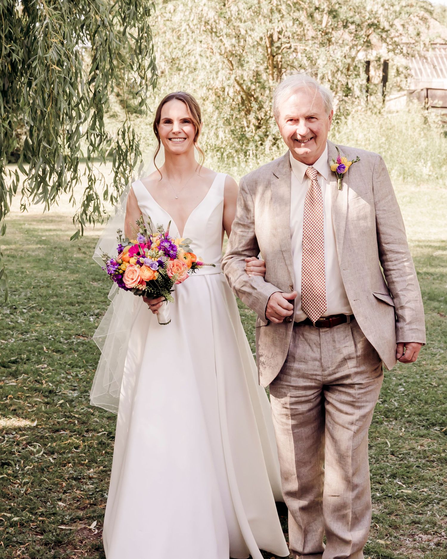 Bride in white gown holding colorful bouquet walks arm-in-arm with older man in tan suit at outdoor wedding.