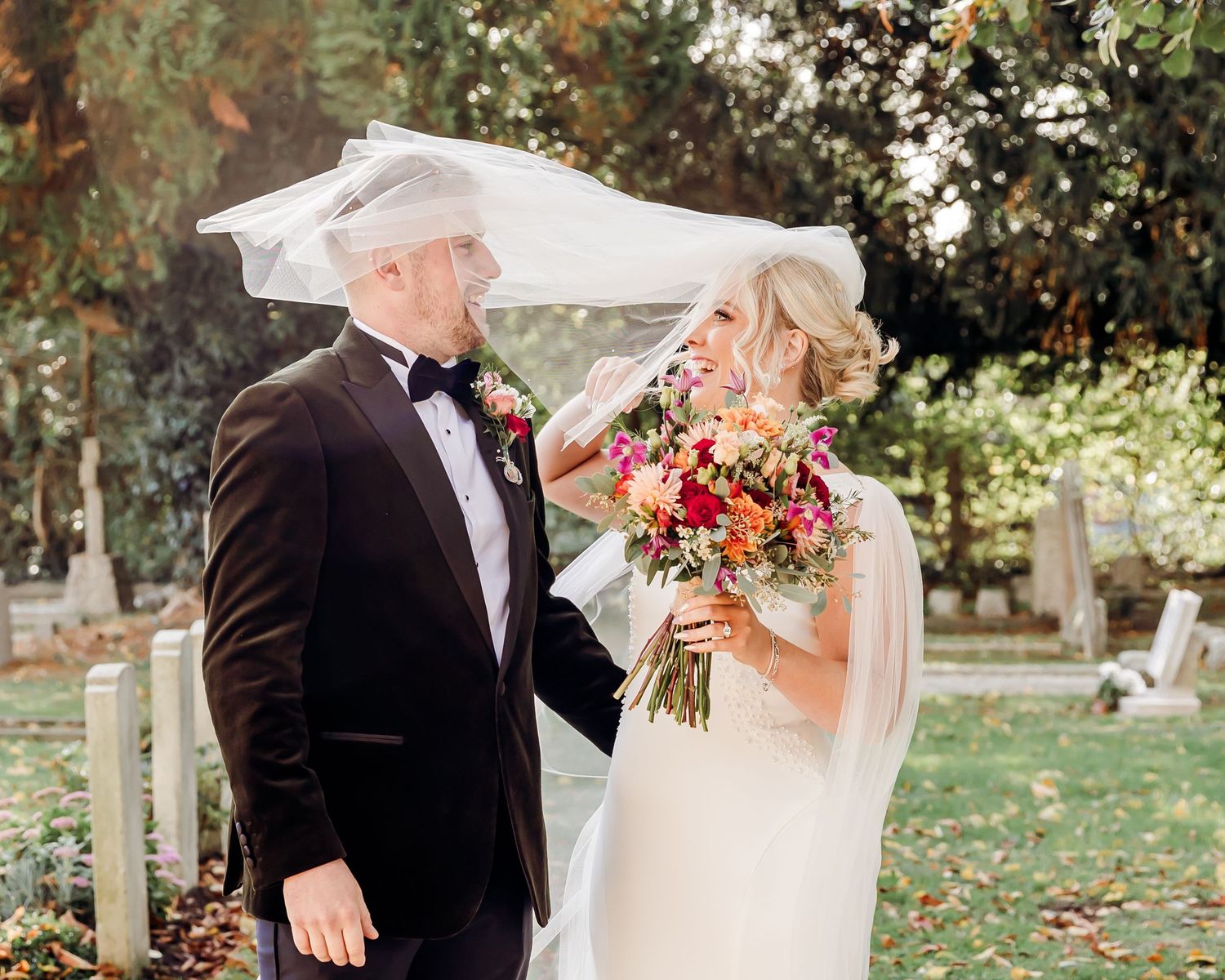 Bride and groom laugh as wind lifts the bridal veil in a sunlit churchyard with autumn foliage.