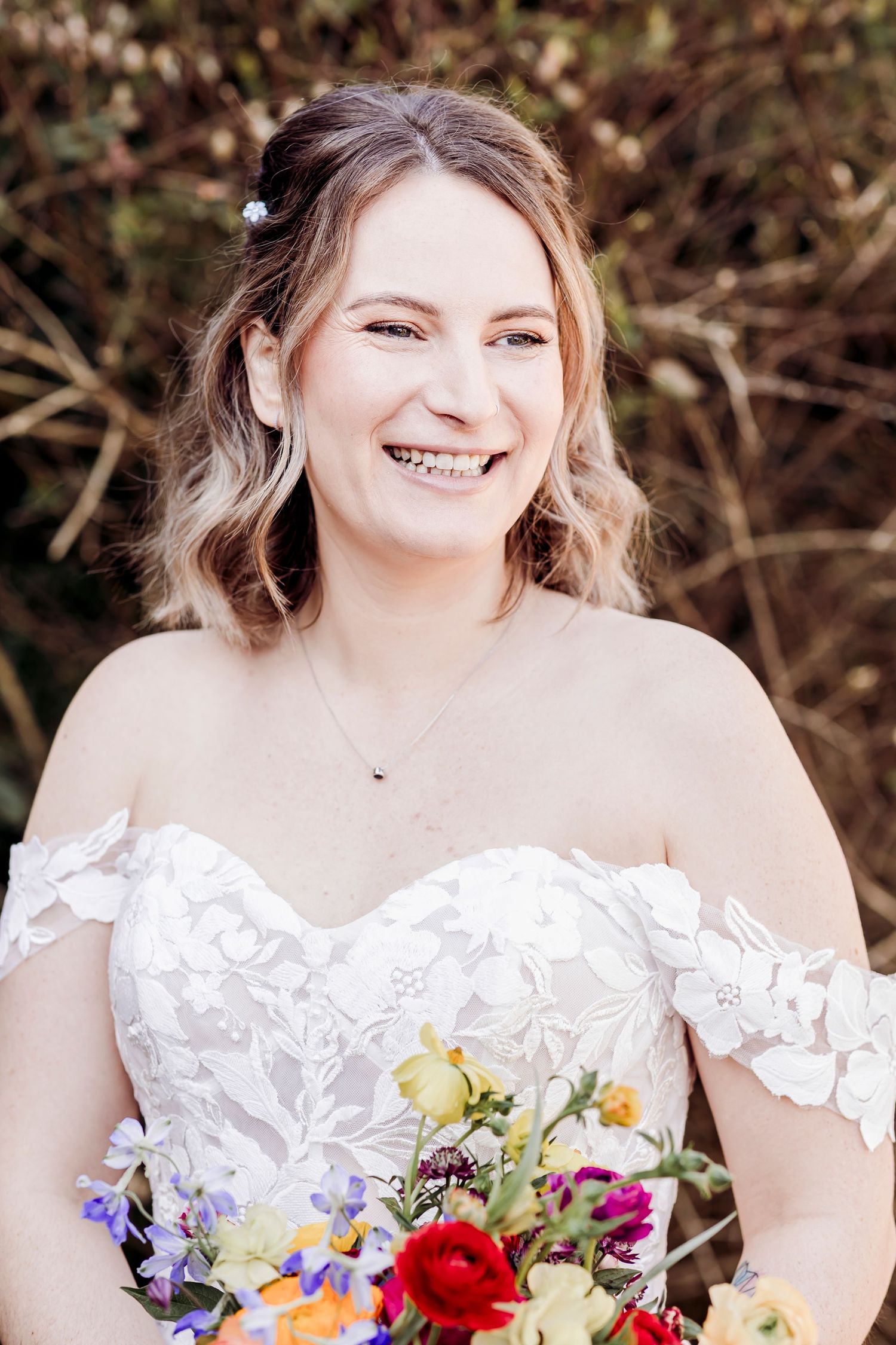 Smiling bride in off-shoulder lace wedding gown holds vibrant colorful bouquet outdoors on her wedding day.