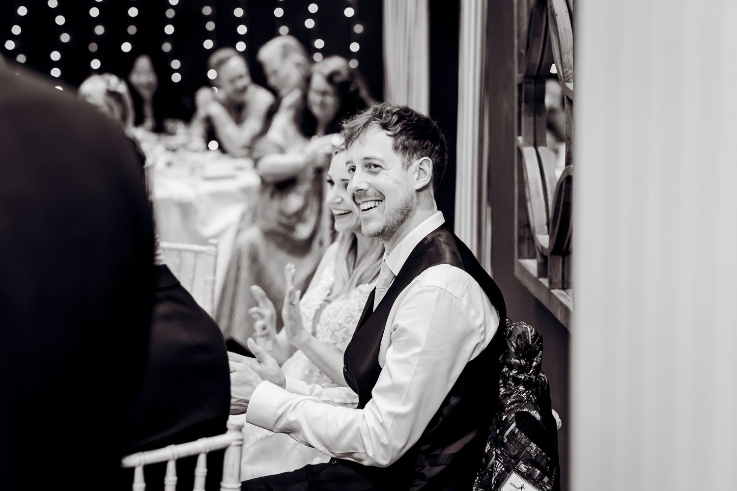Black and white photo of a smiling groom watching wedding speeches with a guest beside him.