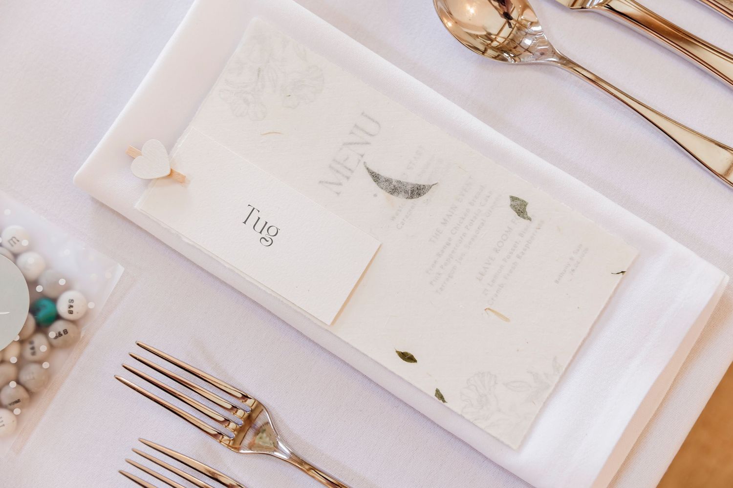 Elegant wedding place setting with gold cutlery, menu card, and pearl bracelet on white tablecloth.
