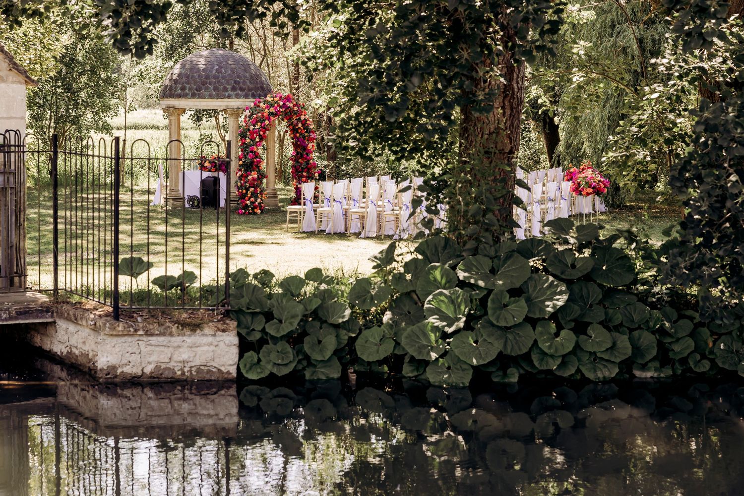 Elegant outdoor wedding ceremony setup with floral arch, white chairs, and gazebo surrounded by lush greenery reflecting in a pond.