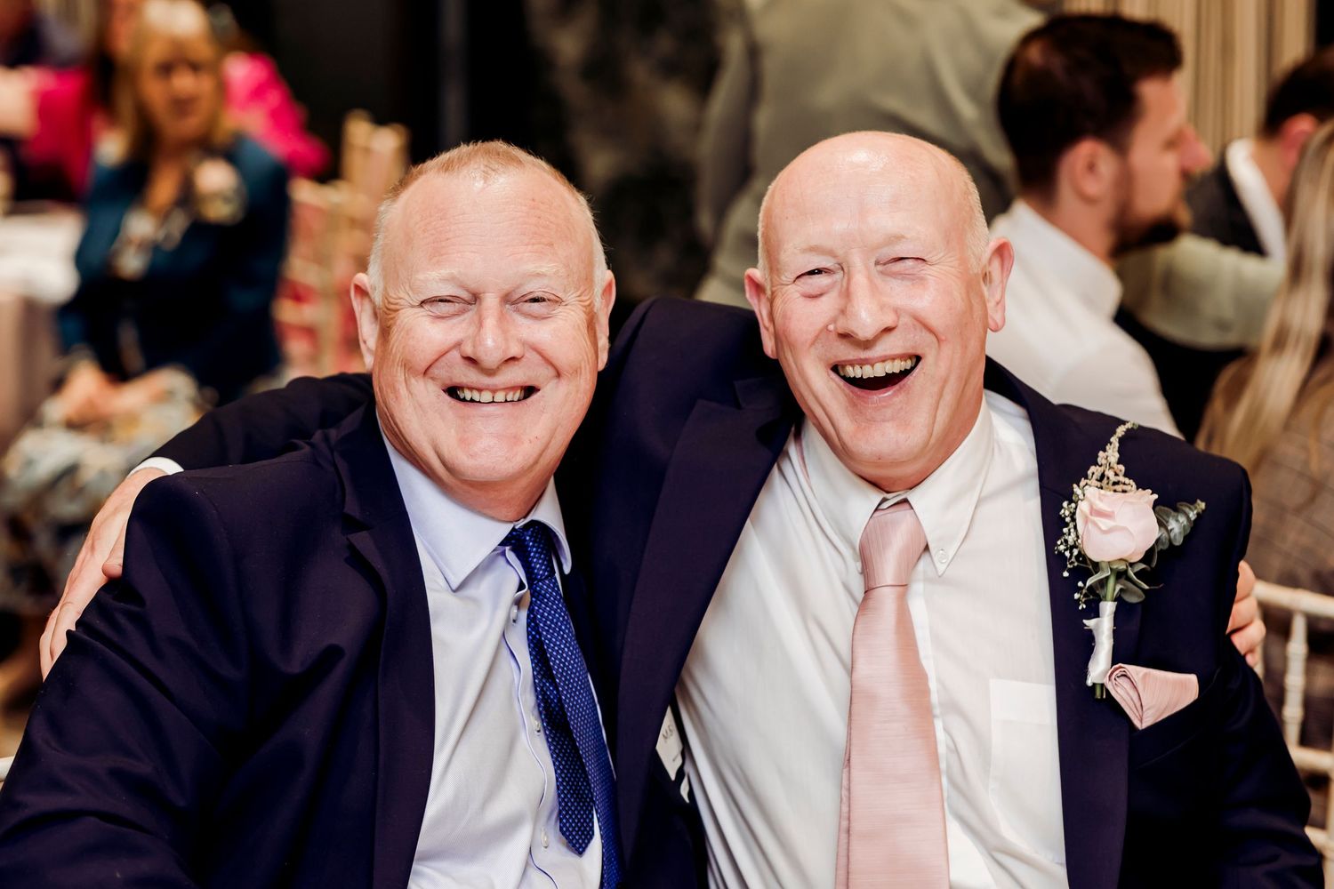 Two cheerful men in navy suits smiling together at a wedding reception celebration.
