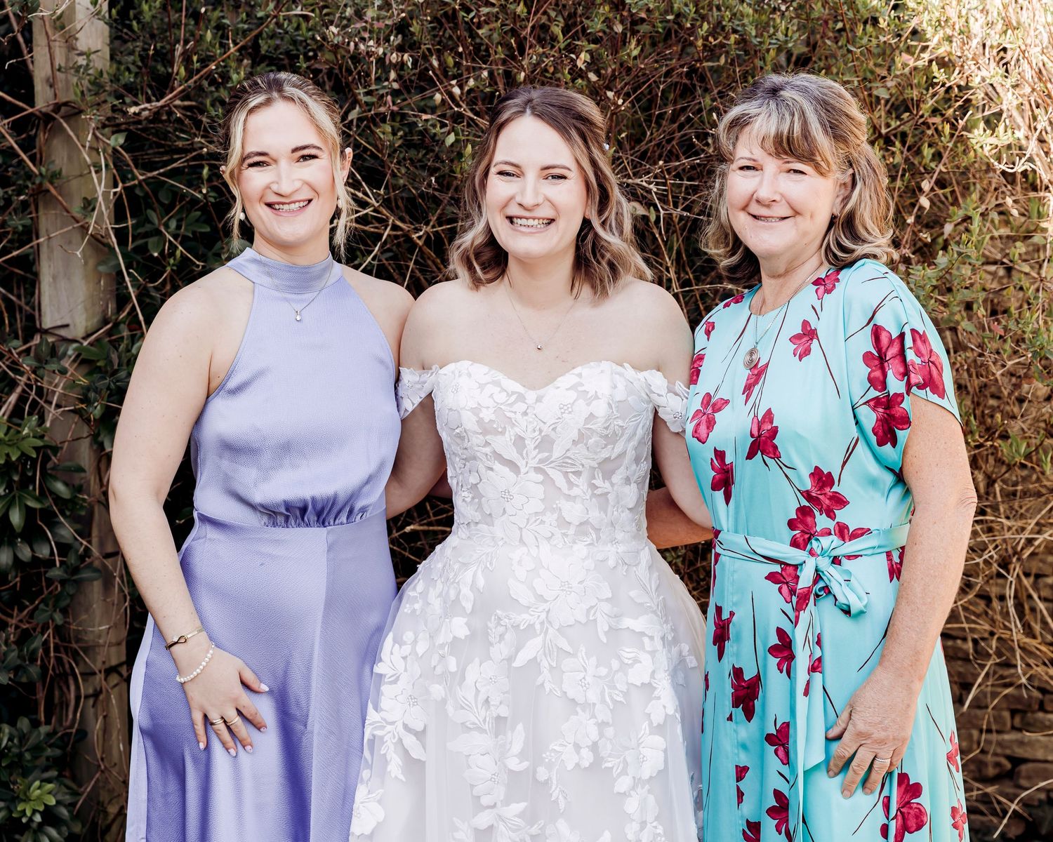 Bride in white lace gown poses with bridesmaid in lavender dress and guest in floral top at outdoor wedding.