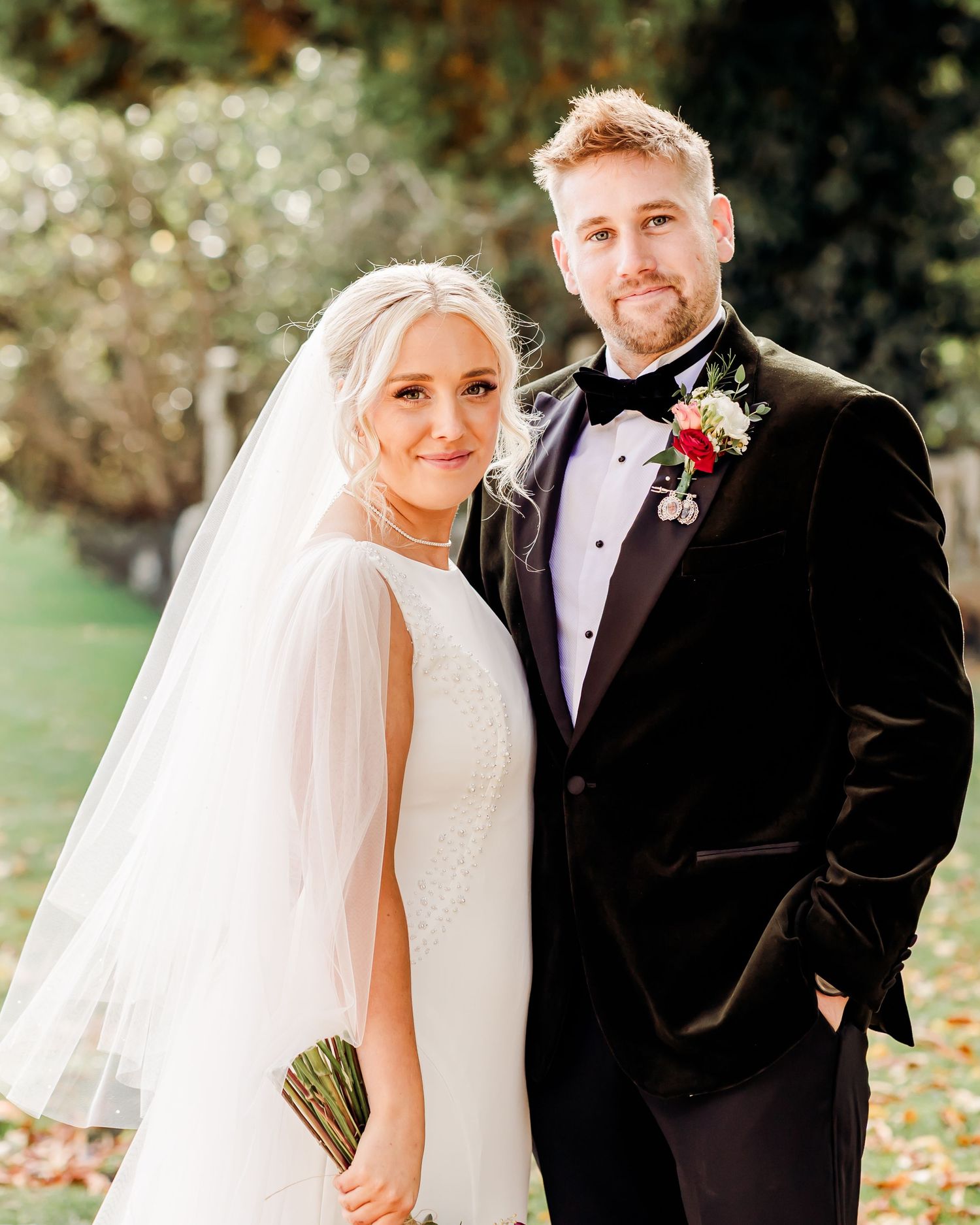 Bride in white gown with veil and groom in black tuxedo pose outdoors at autumn wedding.