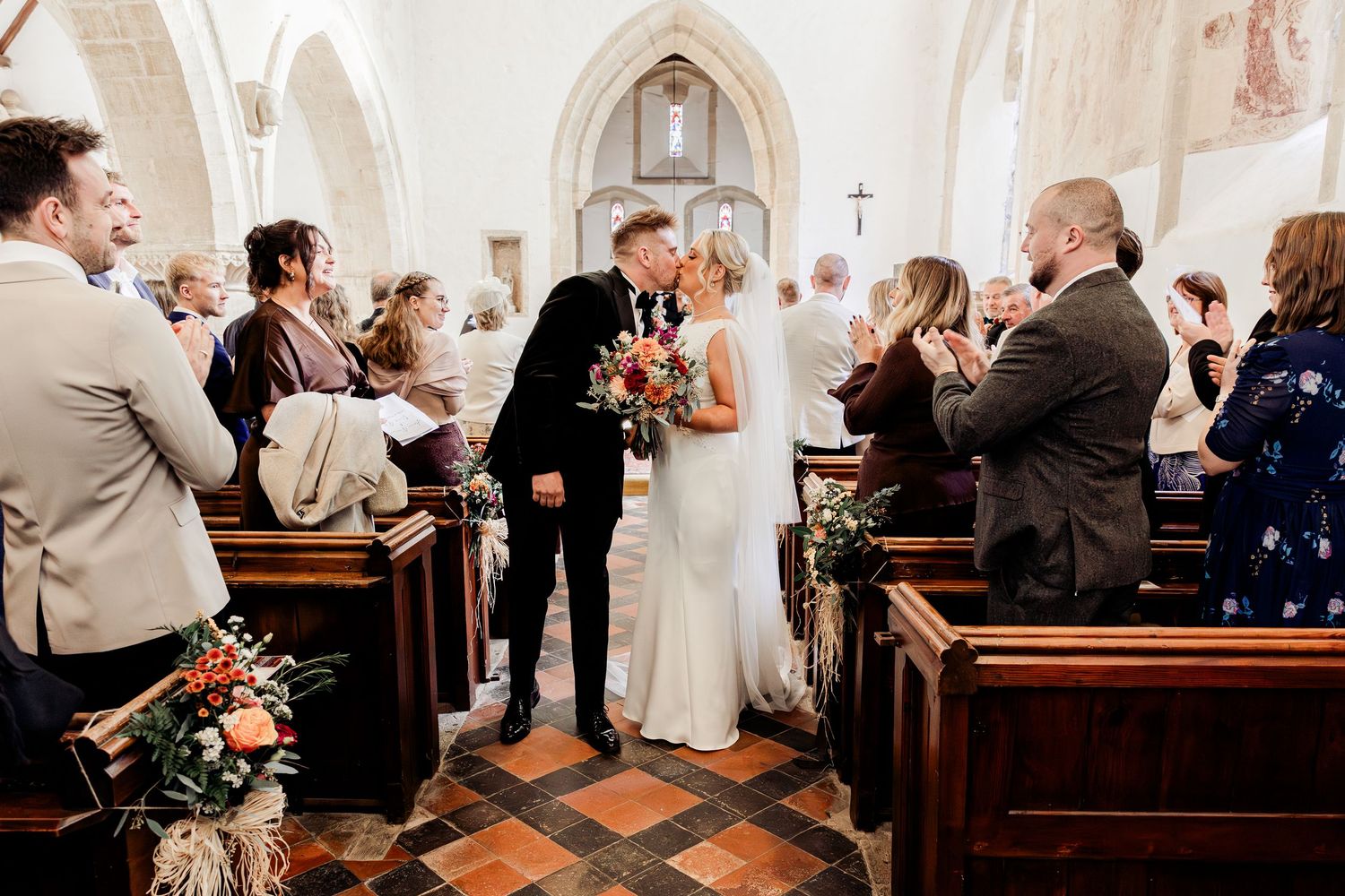 Newlyweds share a kiss in the church aisle surrounded by applauding guests after their ceremony.