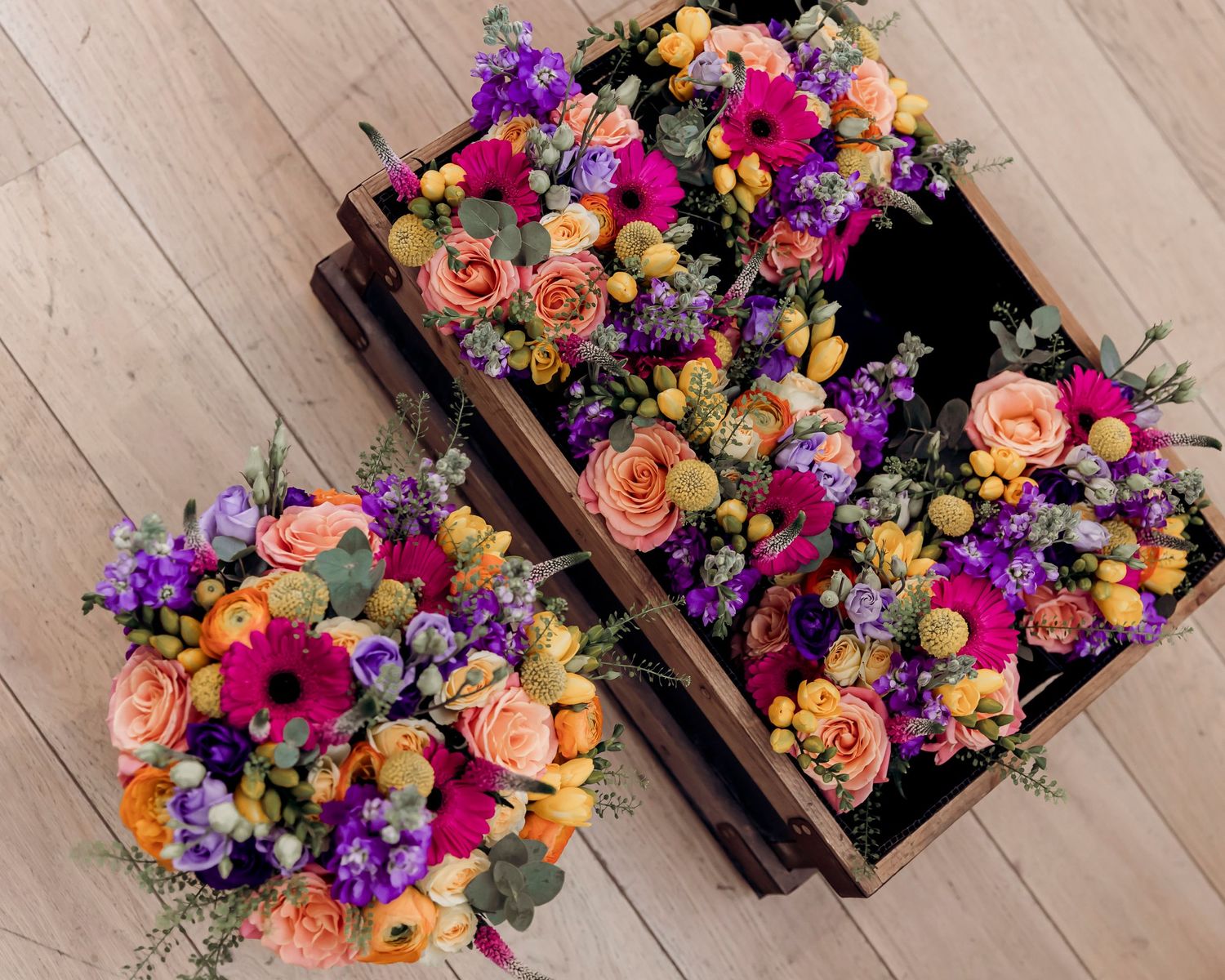 Vibrant mixed flower bouquets with pink, purple, orange roses and gerberas arranged in a rustic wooden crate on wood floor.