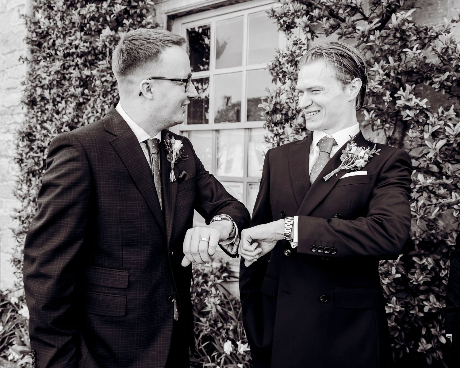Two grooms in dark suits sharing a joyful handshake outside on their wedding day in black and white.