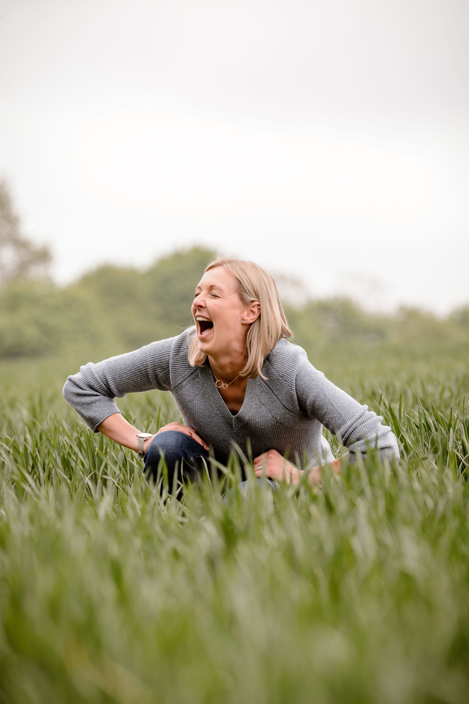 Laughing blonde woman in a grey sweater crouching in a lush green grass field on an overcast day.