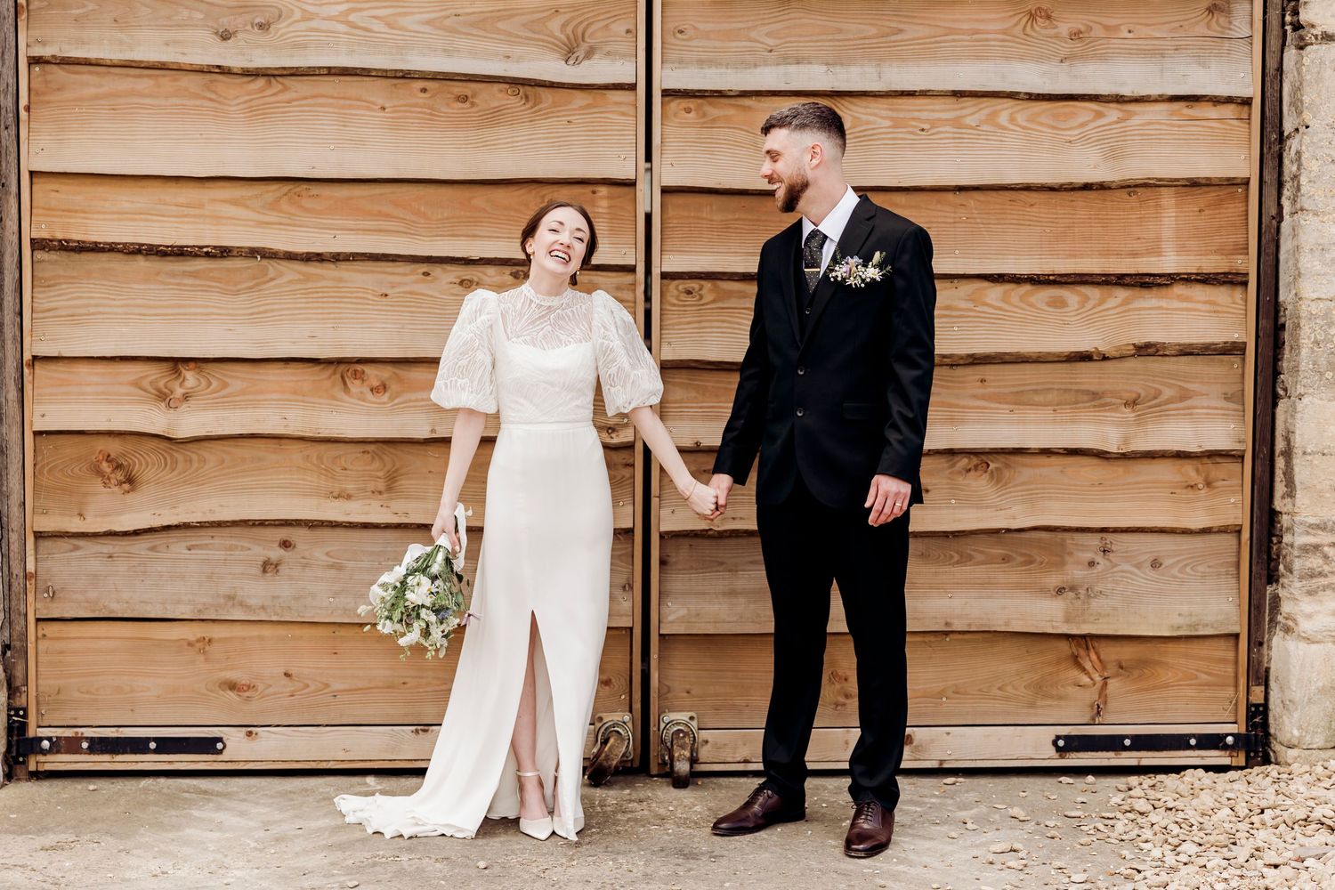 Bride and groom hold hands in front of wooden barn doors, bride holding a wildflower bouquet.