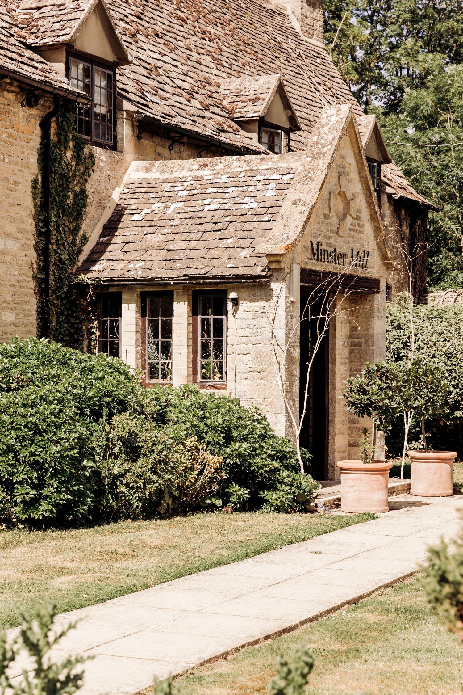 A charming stone Cotswold cottage with lush green gardens and a welcoming entrance sign.