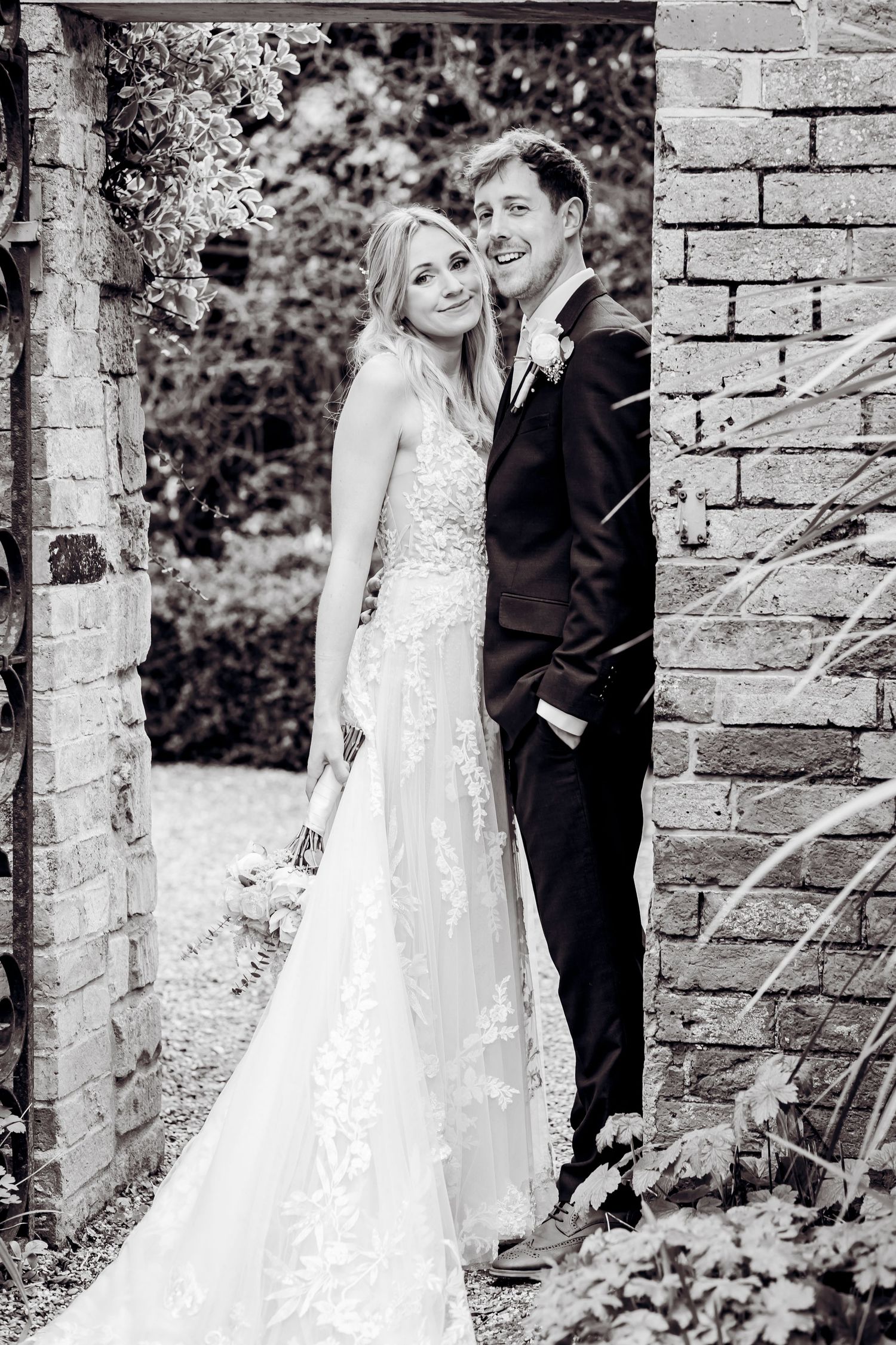 Black and white portrait of bride and groom standing together in a brick archway at their wedding.