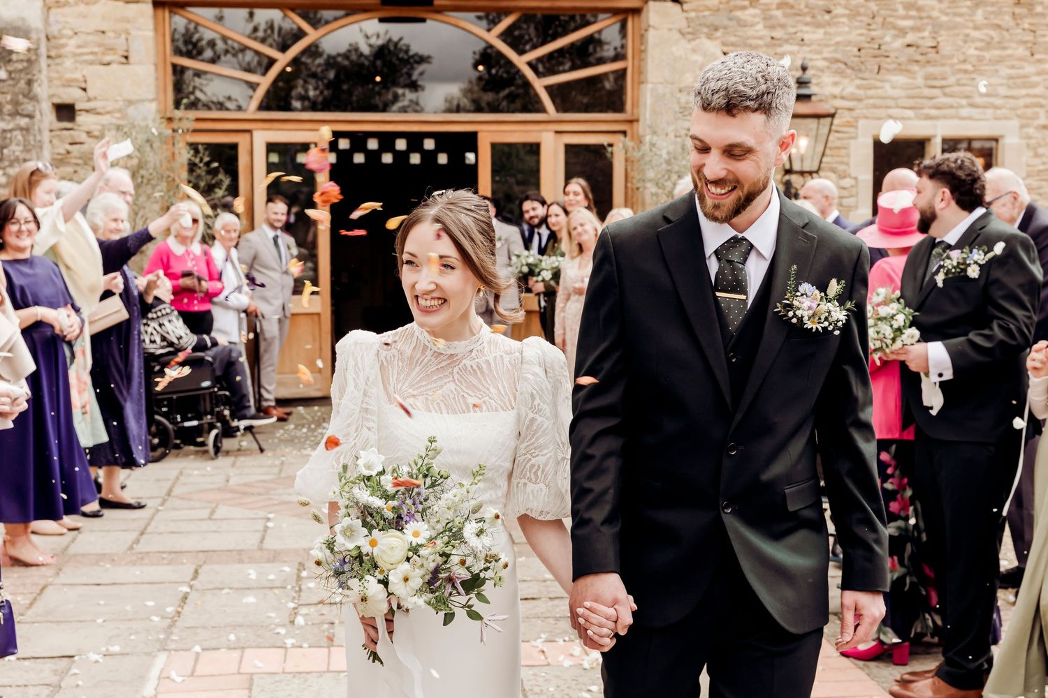 Happy bride and groom exit stone wedding venue holding hands amid guests.