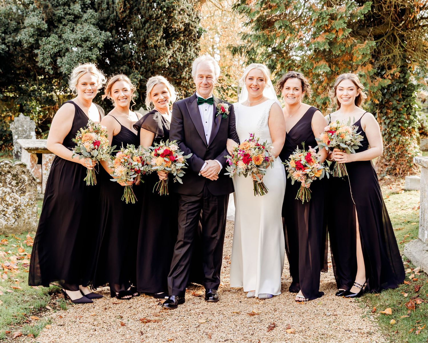 Bride in white gown with groom and four bridesmaids in black dresses holding autumn bouquets outdoors.
