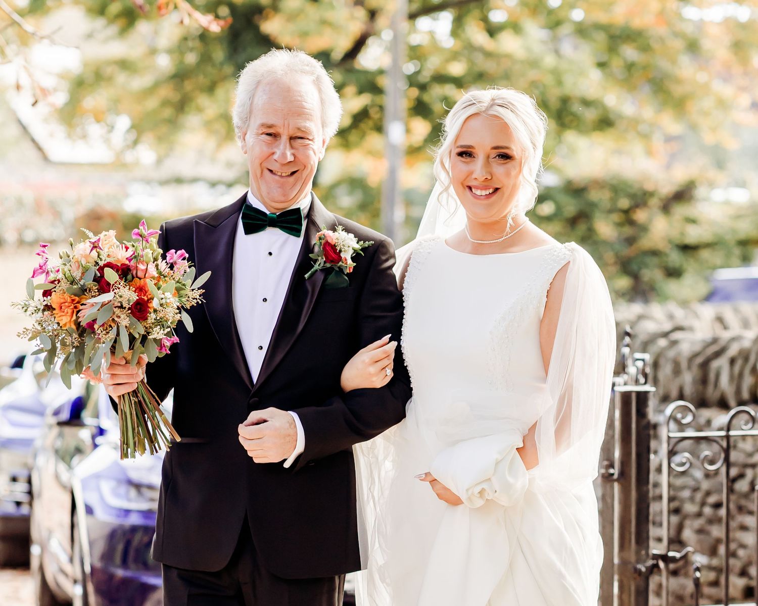 Smiling bride in elegant white gown walking with father holding colorful bridal bouquet outdoors.