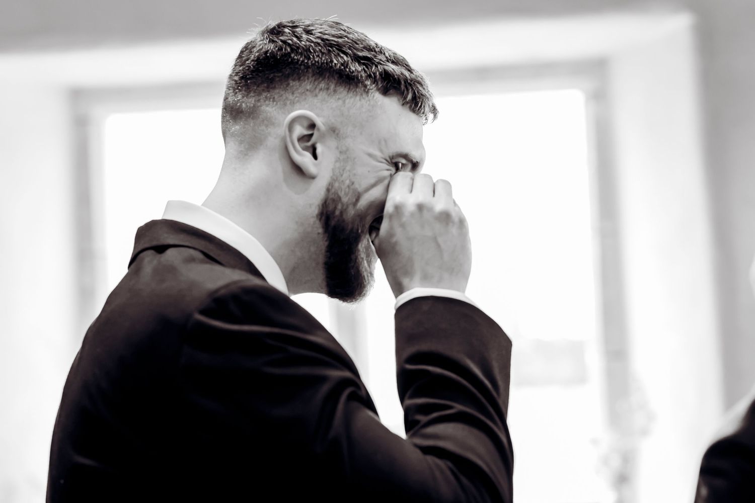 Emotional groom in black suit wiping tears during wedding ceremony, captured in black and white photography.