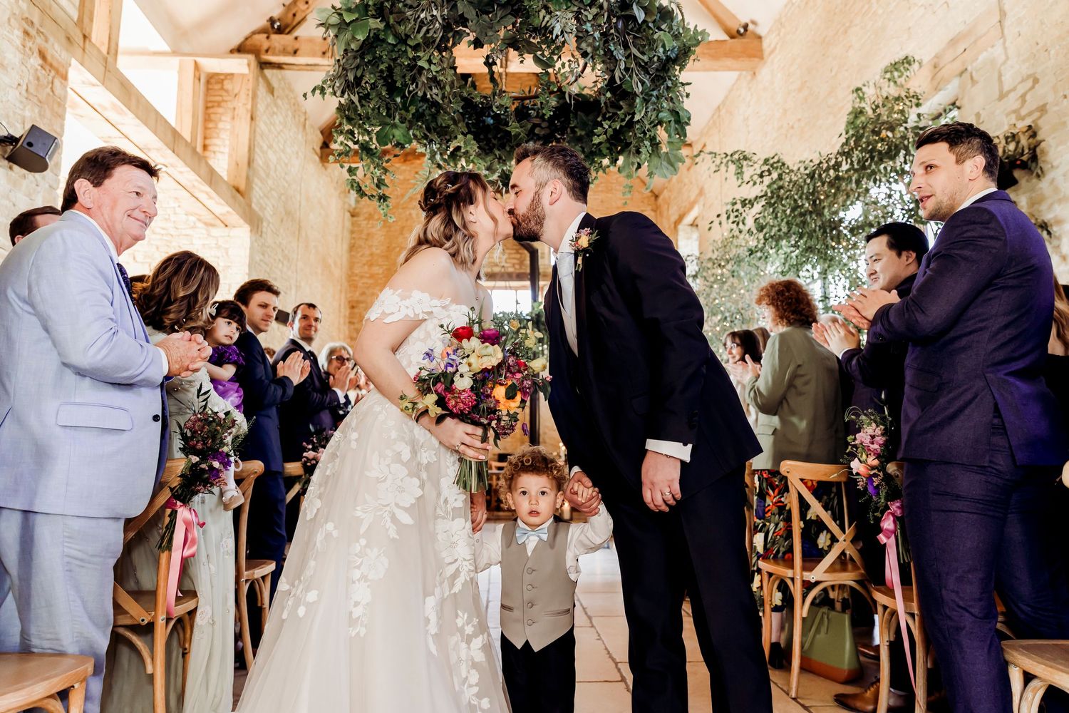 Newlyweds kiss at altar with young boy and applauding guests in rustic barn venue.