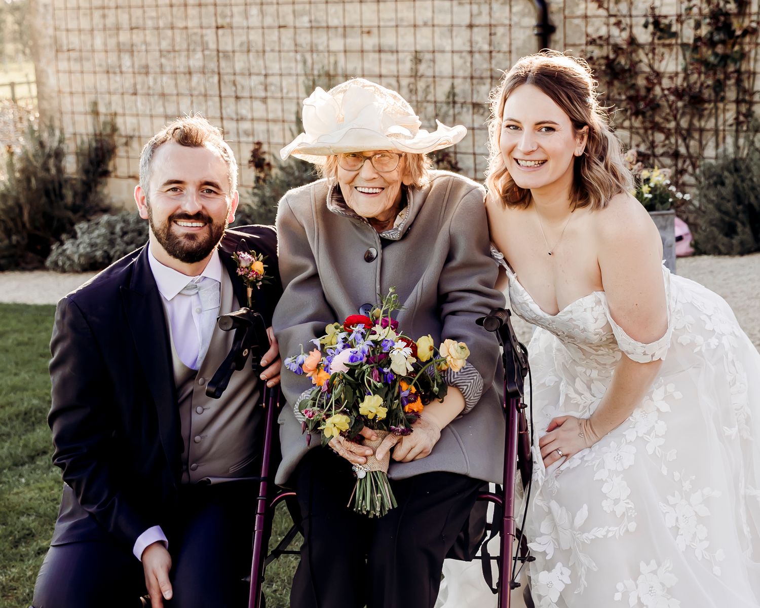 Bride and groom kneel beside elderly grandmother in hat holding colorful bridal bouquet in garden setting.