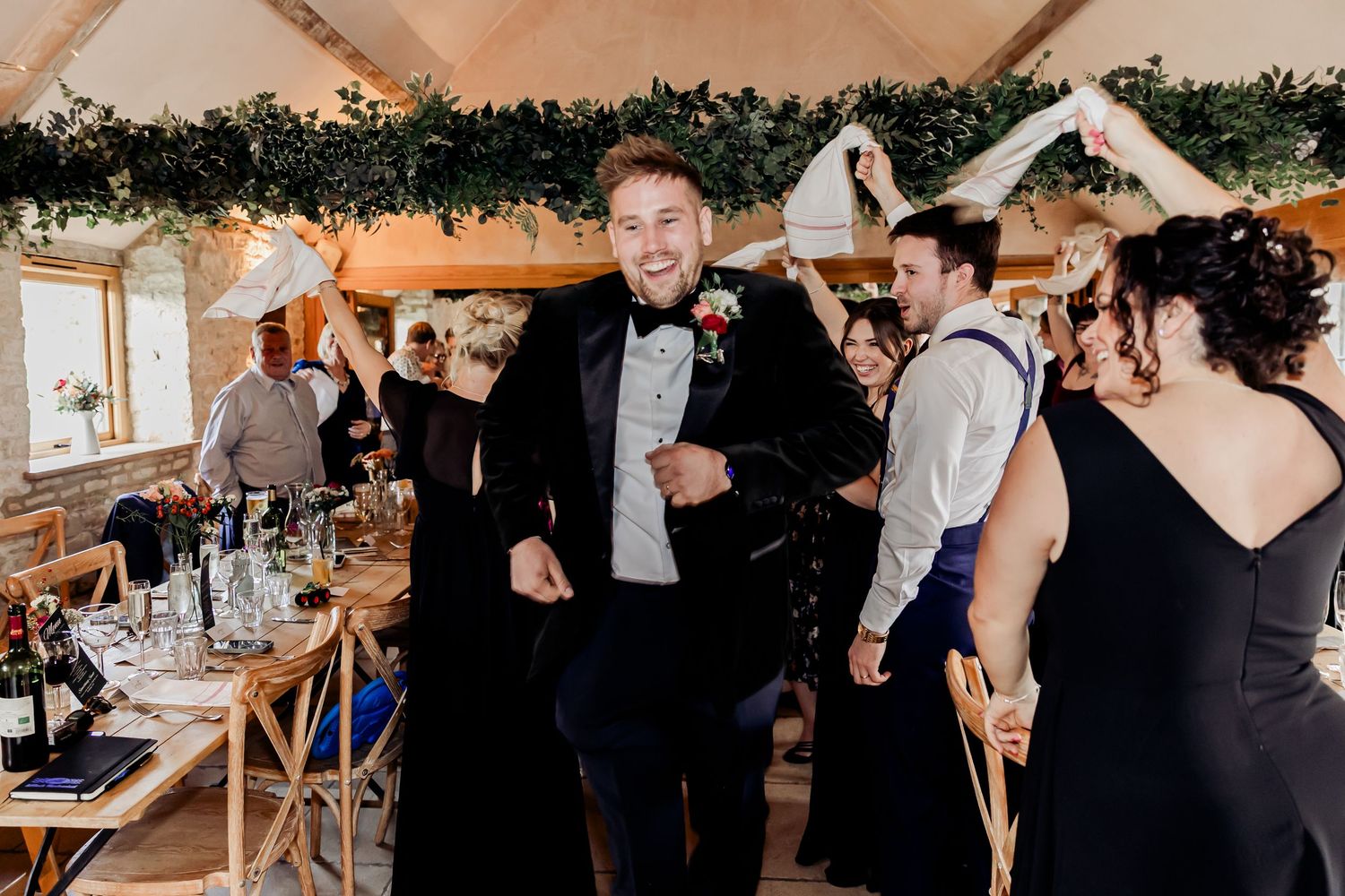 Groom in black tuxedo dancing and celebrating energetically with wedding guests in a rustic barn reception.