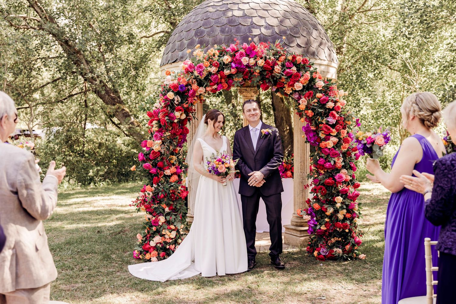 Bride and groom stand at colorful floral arch ceremony backdrop outdoors surrounded by wedding guests in lush garden setting.