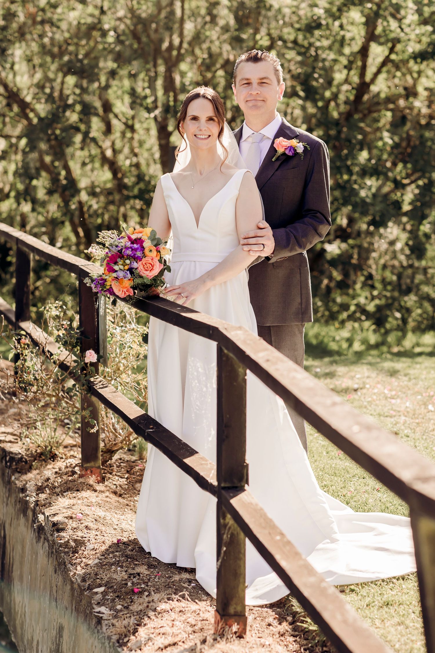 Bride and groom pose on wooden bridge surrounded by lush greenery holding vibrant wedding bouquet.