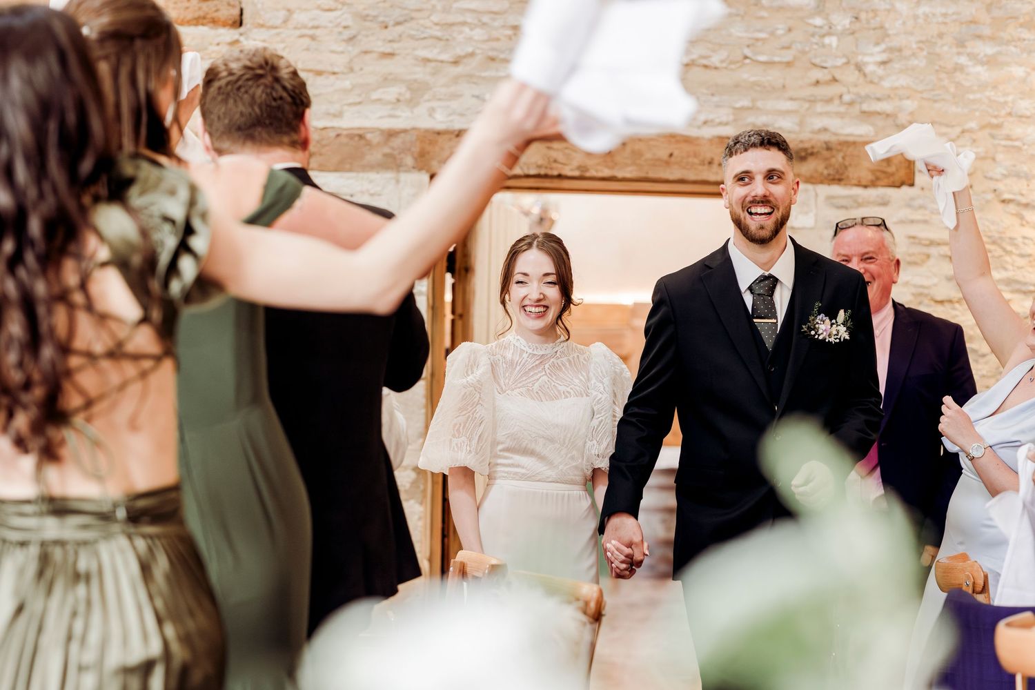 Newlywed couple walks hand in hand through cheering guests waving white ribbons at rustic barn wedding.