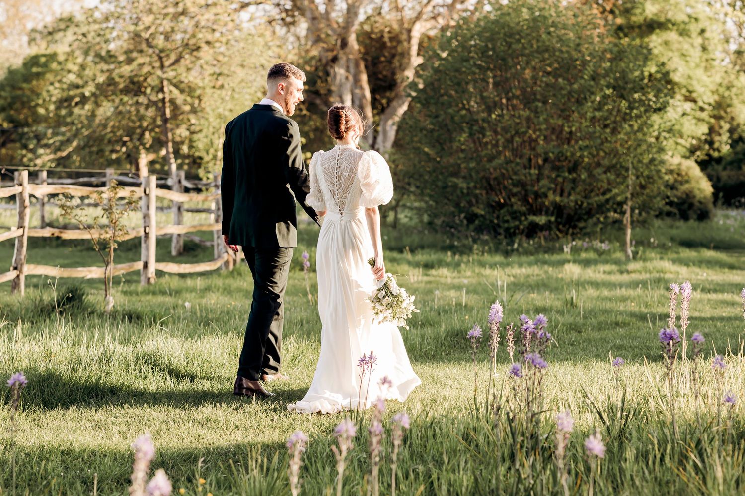 Bride and groom walk hand in hand through a sunlit meadow with purple wildflowers on their wedding day.