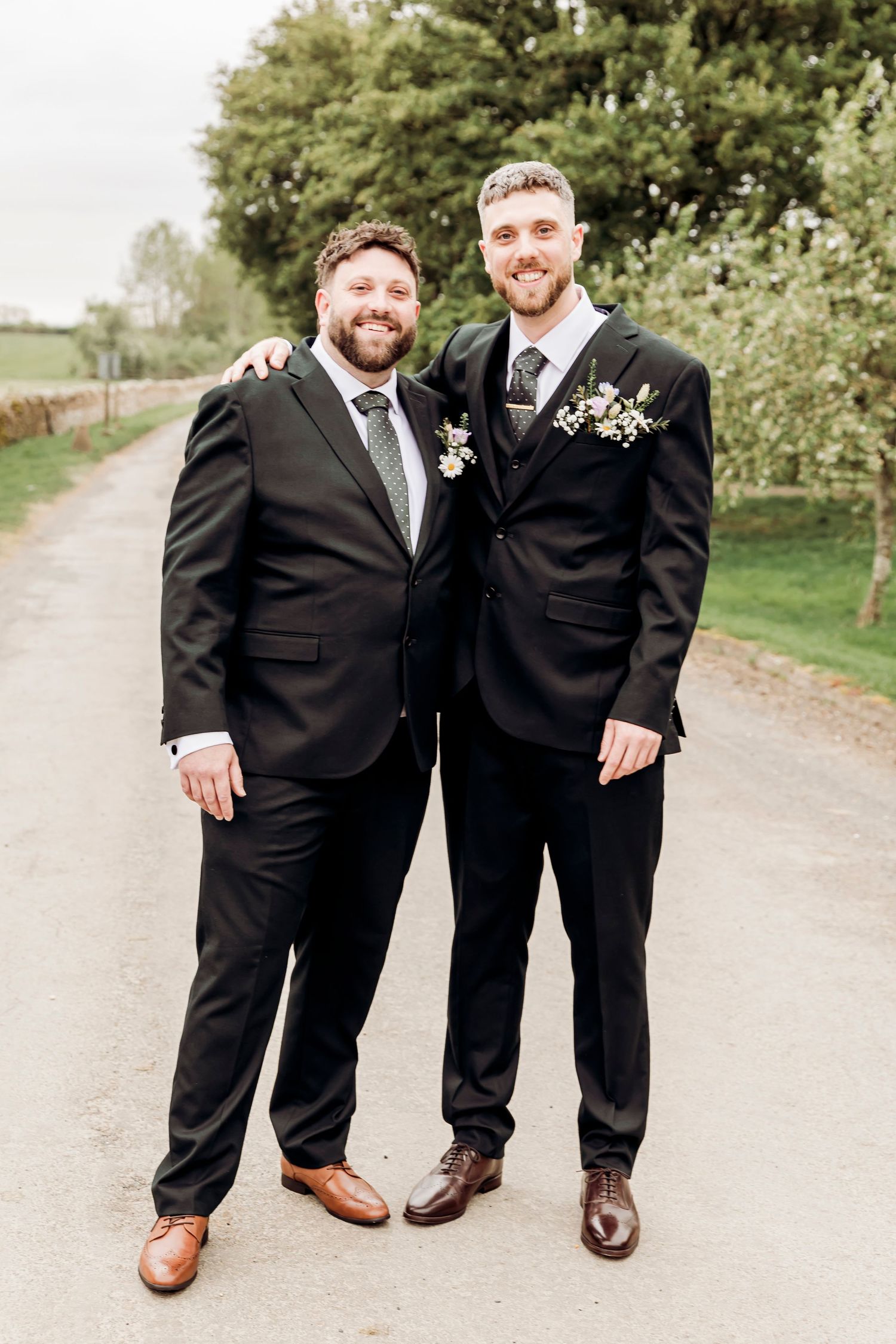 Two groomsmen in black suits with boutonnieres pose together on a tree-lined path at a wedding.