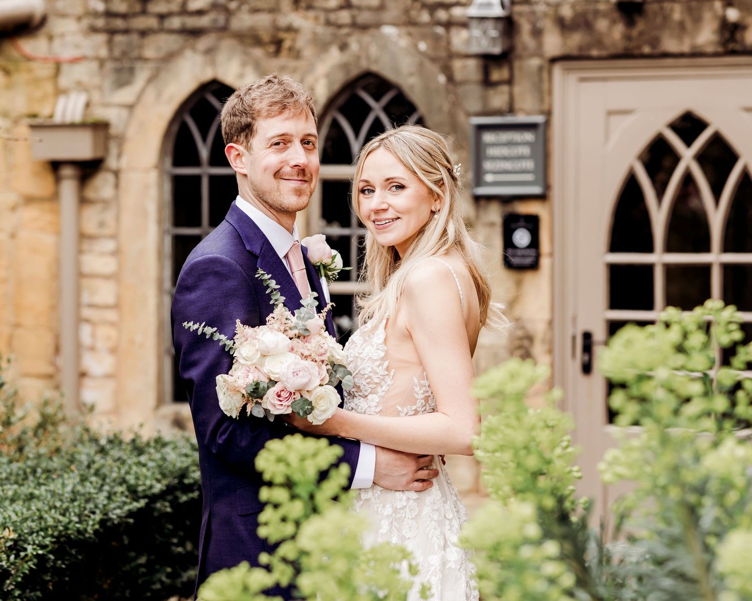 Smiling bride and groom posing together outside a stone church, bride holding a pink floral bouquet.