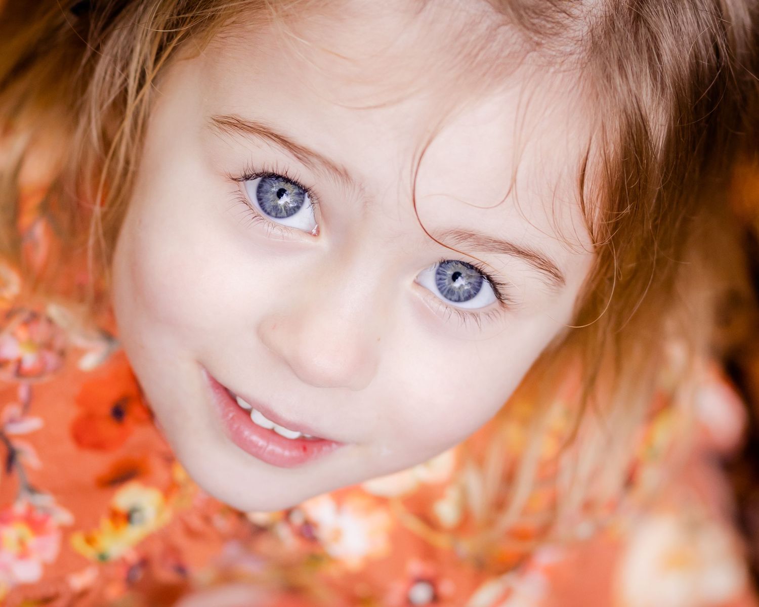 A young girl with blue eyes and auburn hair smiles warmly, wearing an orange floral dress.