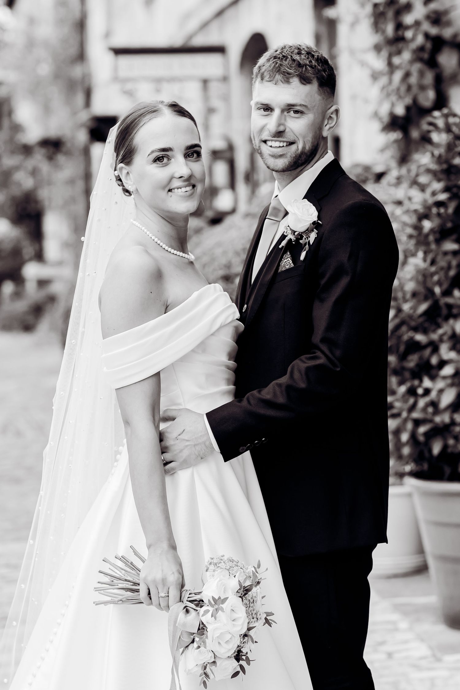 Black and white portrait of smiling bride in off-shoulder gown and groom embracing at outdoor wedding venue.