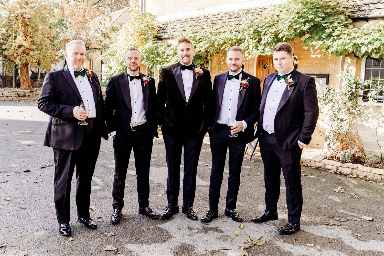Six groomsmen in black tuxedos with green bow ties posing together outdoors at a wedding venue.