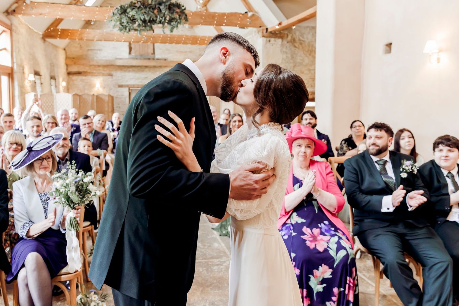 Bride and groom share their first kiss at a rustic barn wedding ceremony with guests watching.