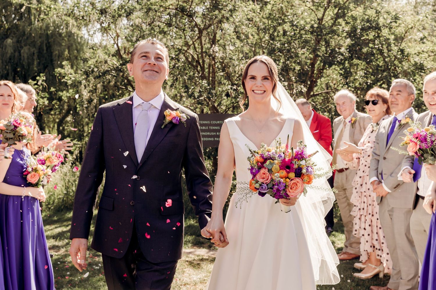 Happy newlyweds walking through confetti shower holding hands with guests celebrating in sunny outdoor wedding.
