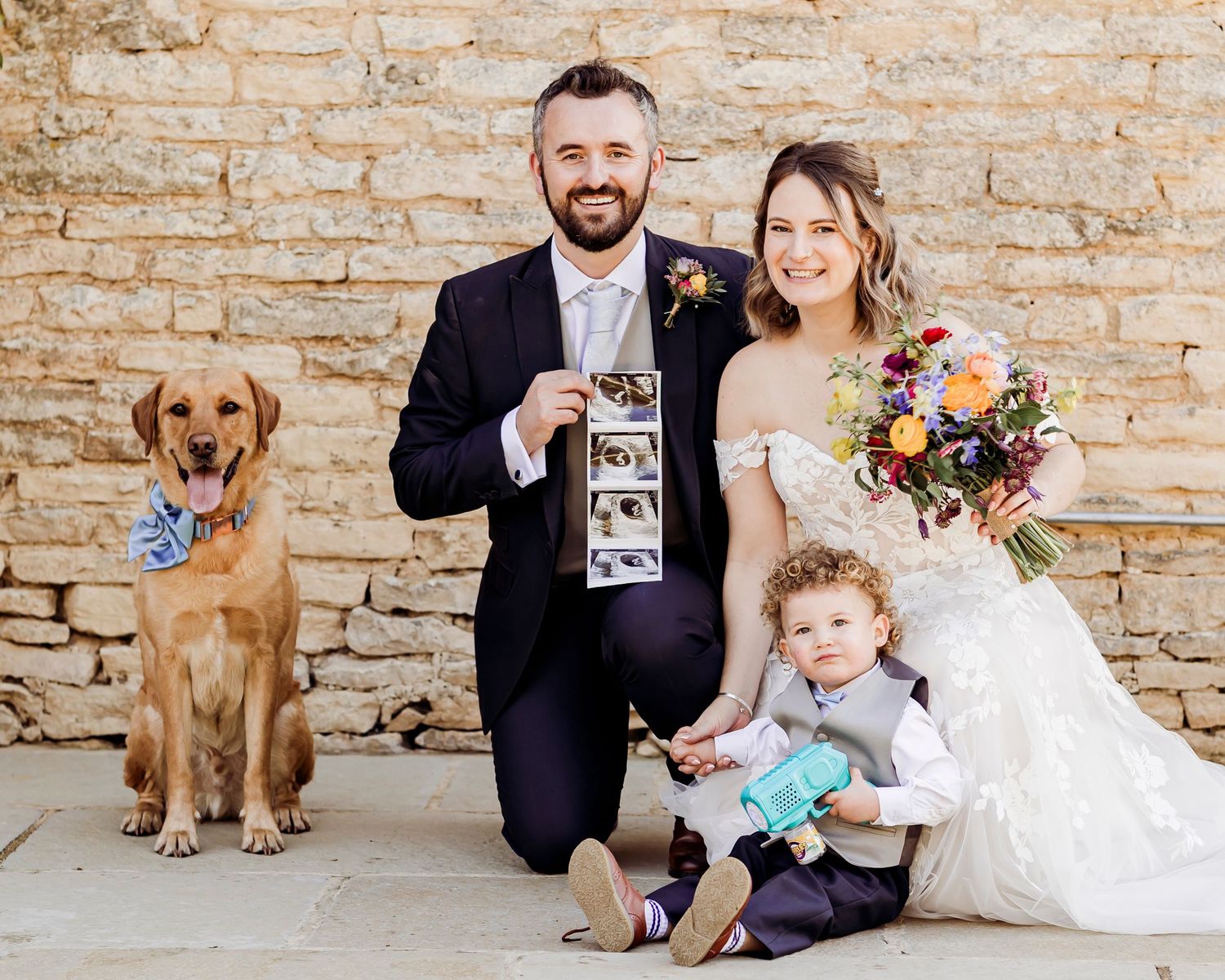 Newlyweds pose with dog, toddler, and ultrasound photos against rustic stone wall at wedding.