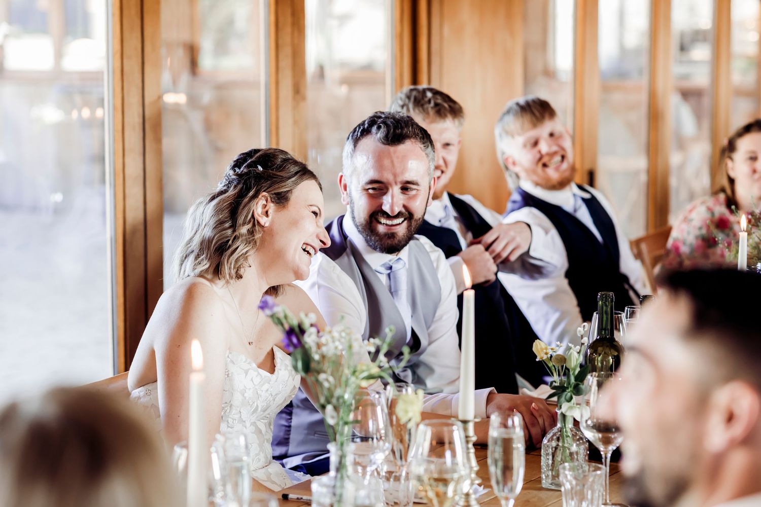 Laughing bride and bearded groom enjoy wedding reception speeches seated at elegantly decorated head table.