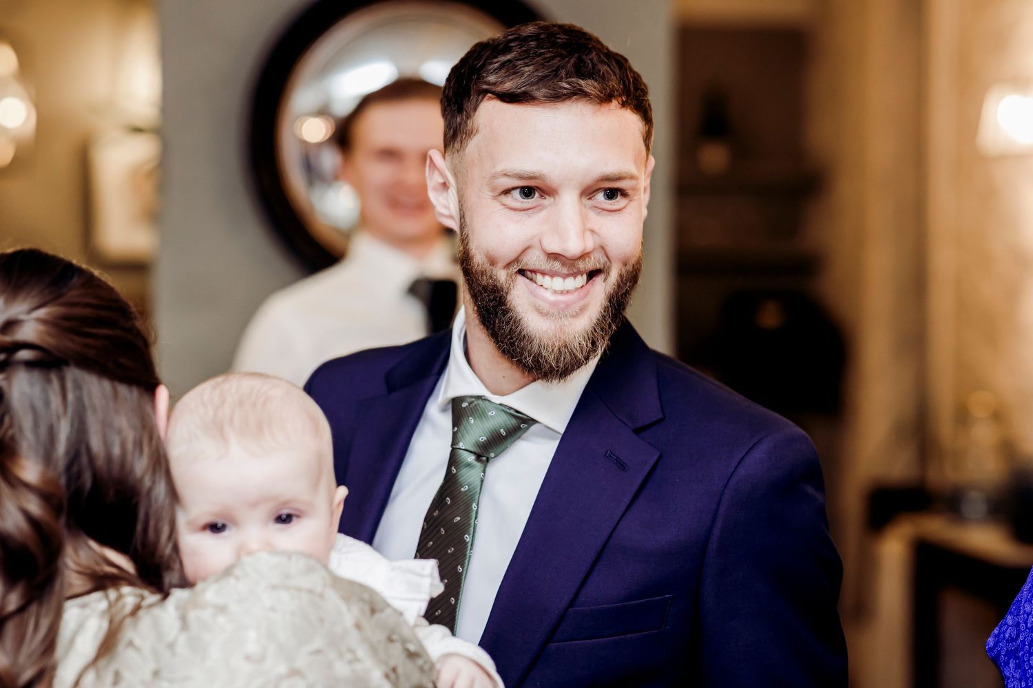 Smiling bearded man in navy suit holding a baby at a wedding reception.