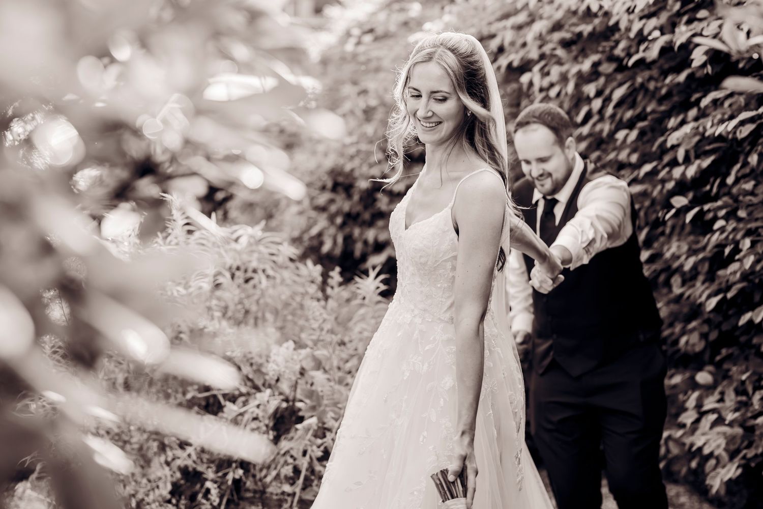 Black and white photo of a smiling bride in lace gown holding hands with groom in suit among lush greenery.