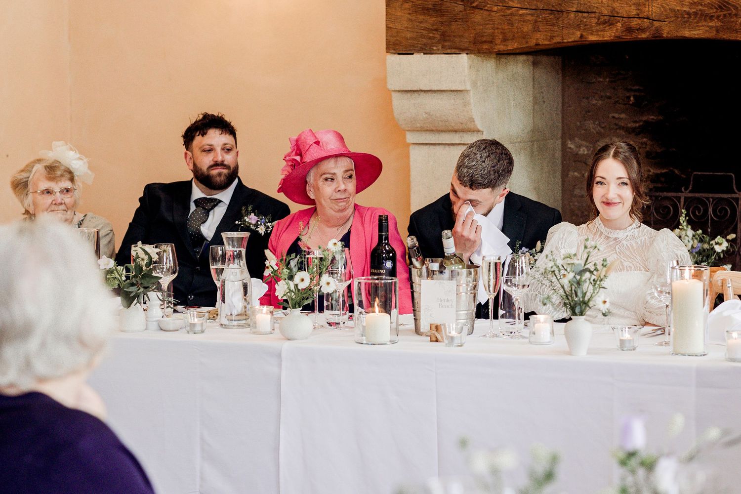 Wedding guests seated at head table with woman in pink hat and suit during indoor reception ceremony.
