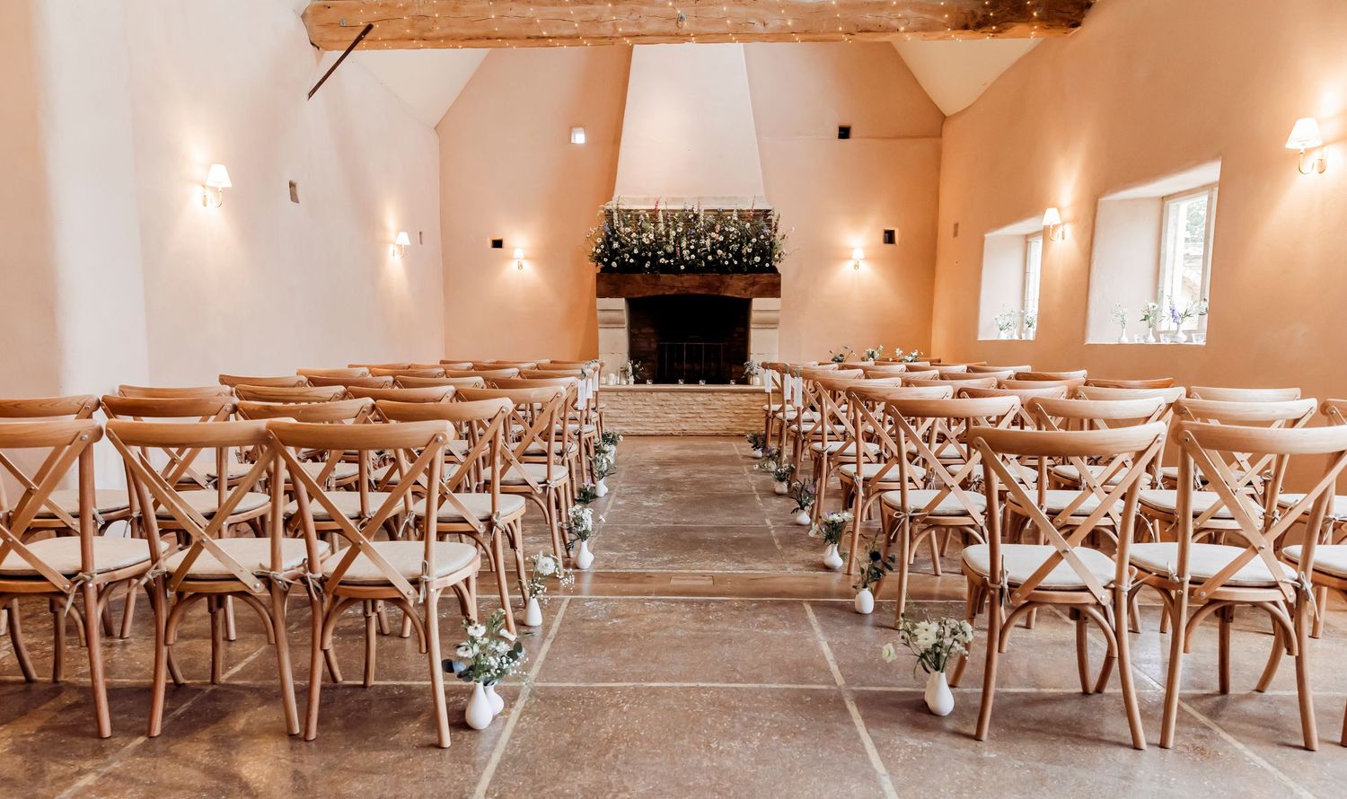 Elegant barn wedding ceremony venue with wooden cross-back chairs and floral aisle decorations.