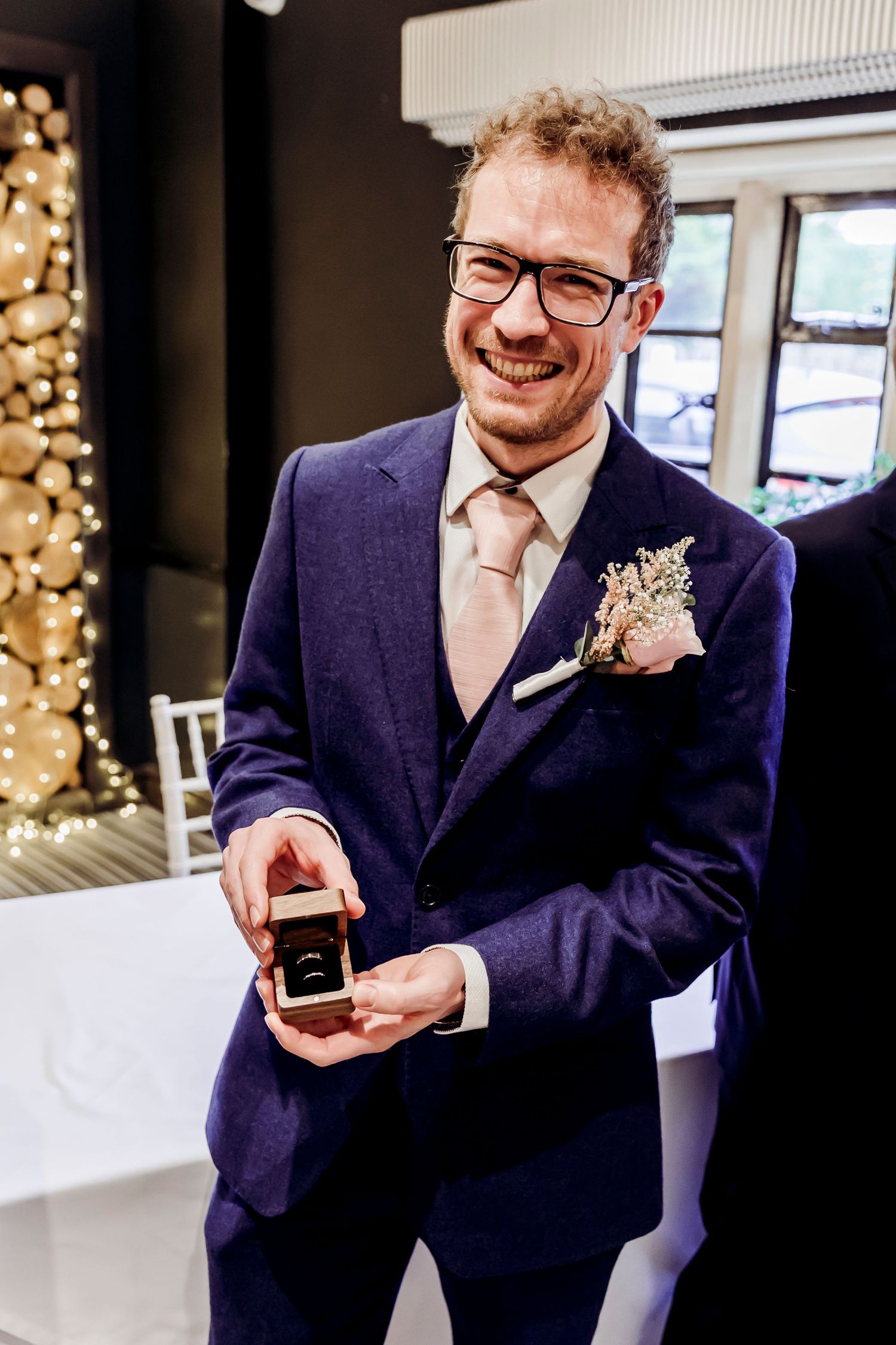 Smiling groom in navy suit presenting wedding ring box with boutonniere at indoor ceremony.