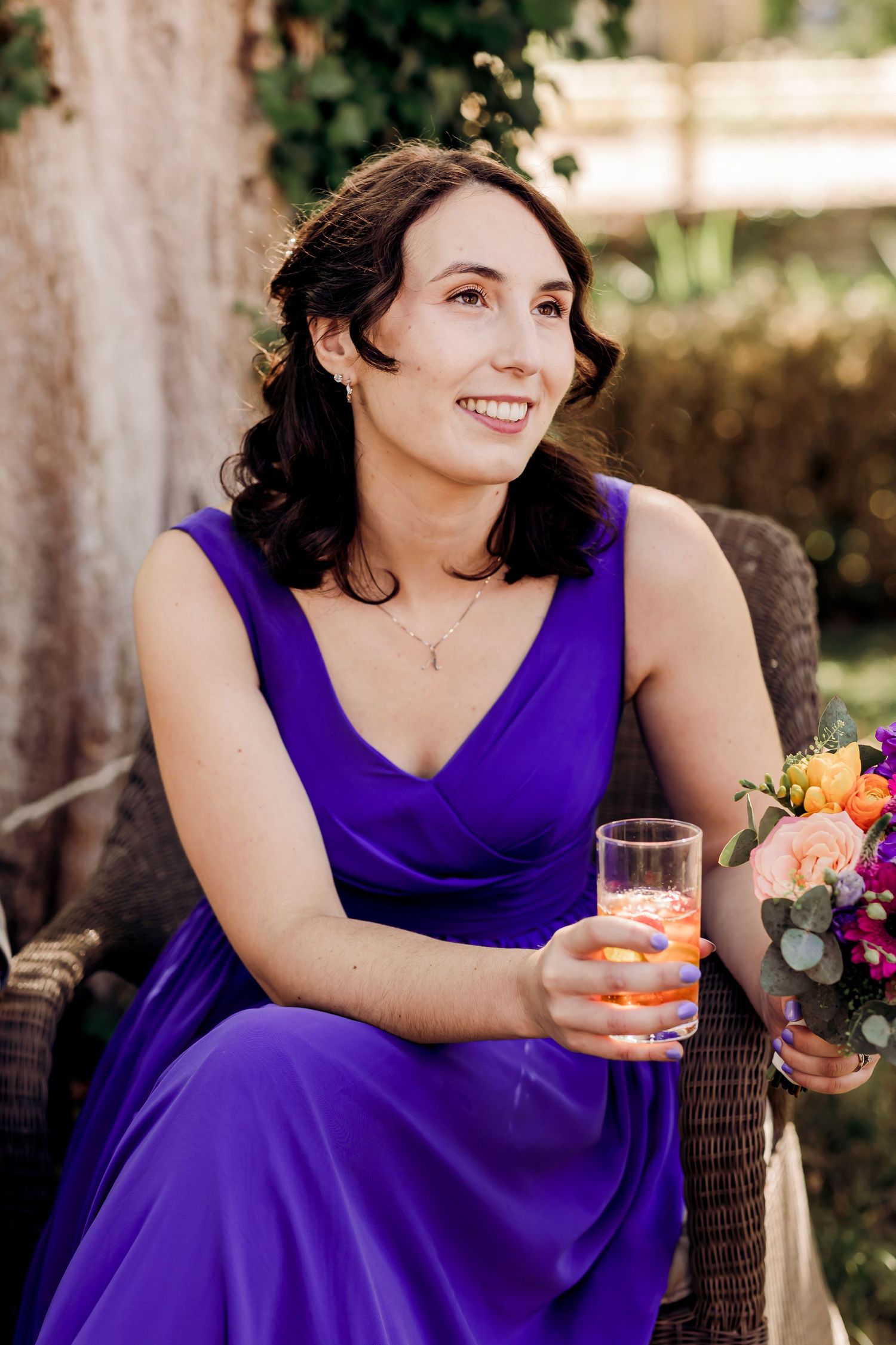Bridesmaid in purple gown smiles while holding drink and colorful bouquet at outdoor wedding reception.