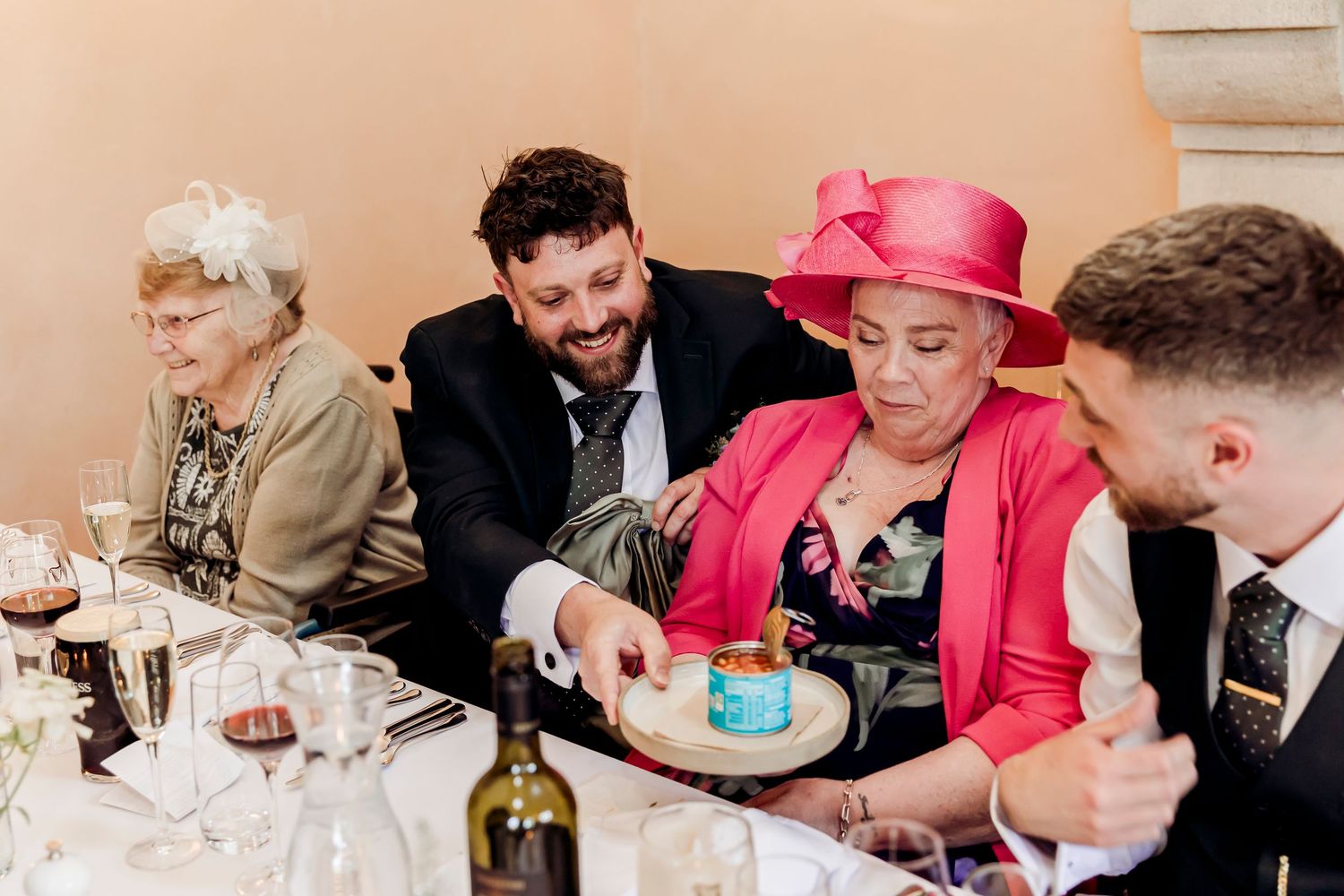 Wedding guests laughing and sharing food at a reception table with wine glasses and bottles.