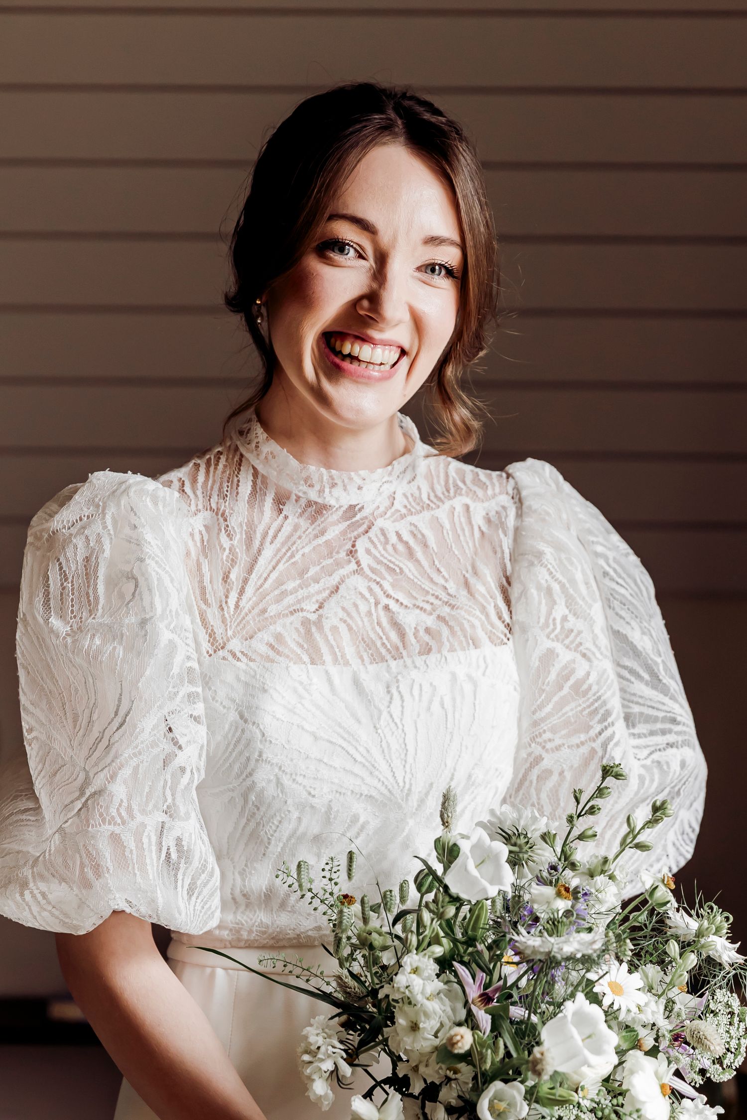 Smiling bride in white lace dress with puff sleeves holding a wildflower bouquet.