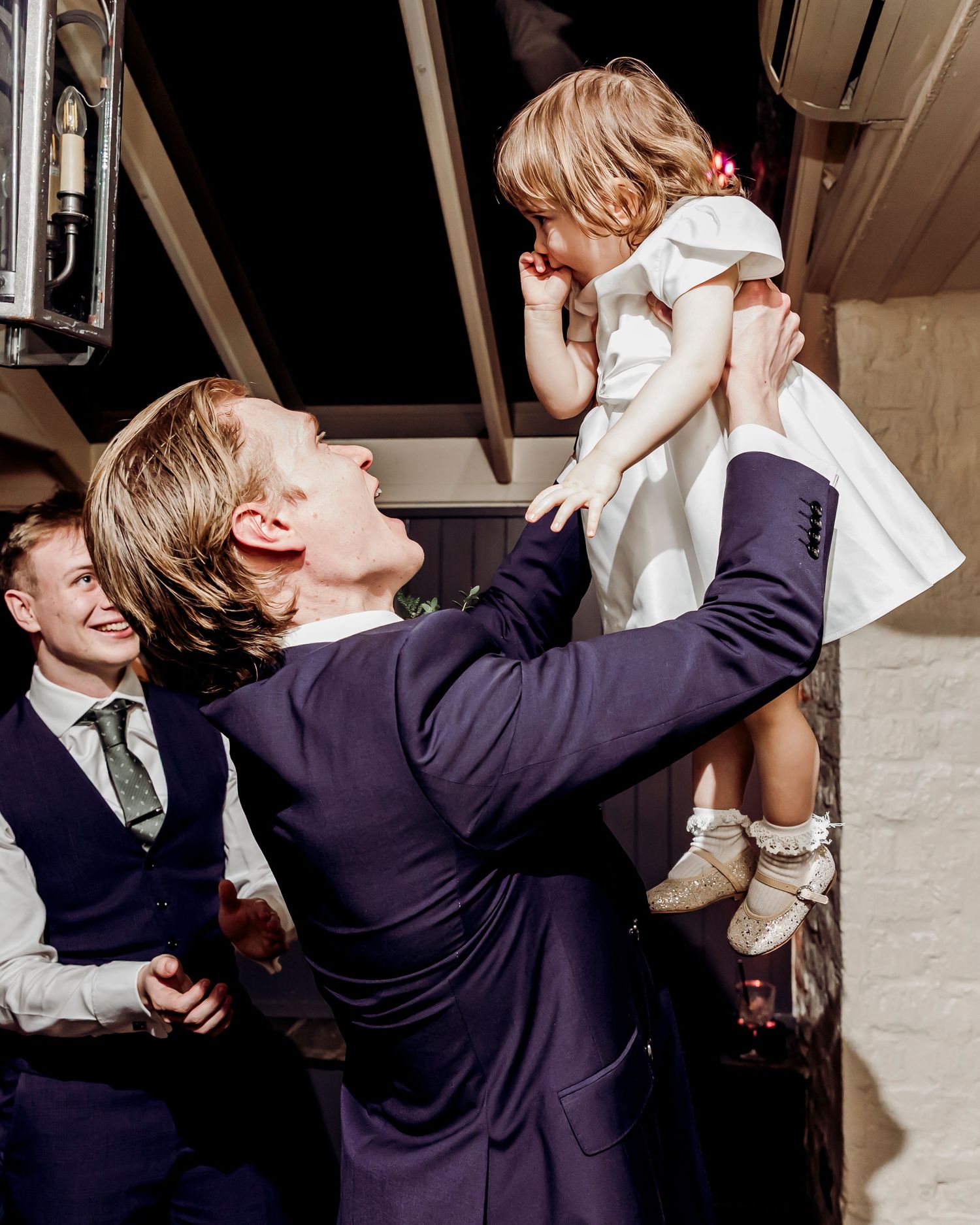 Groom in navy suit joyfully lifting a young flower girl at wedding reception.