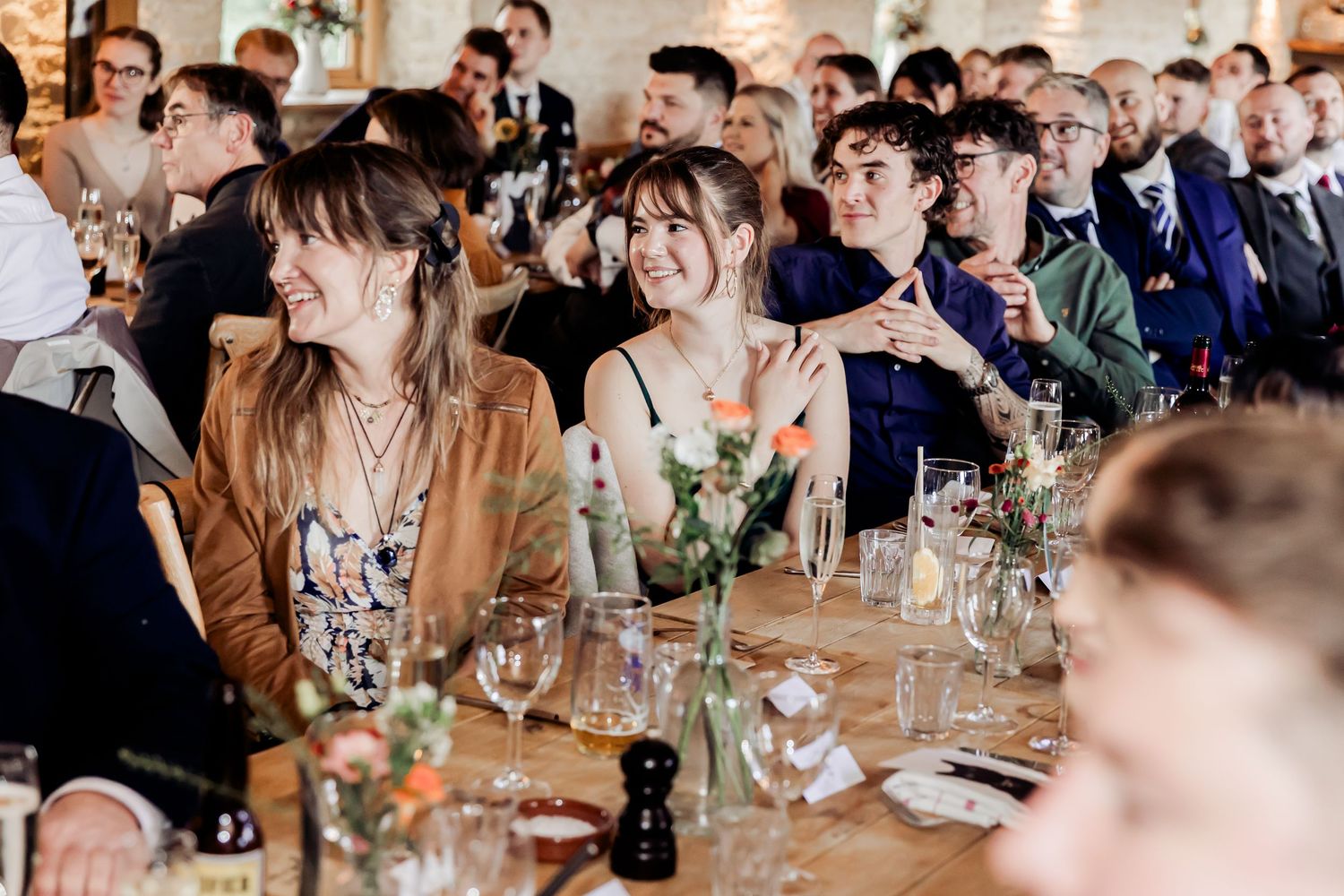Smiling wedding guests enjoy speeches at a rustic barn reception with floral centerpieces.