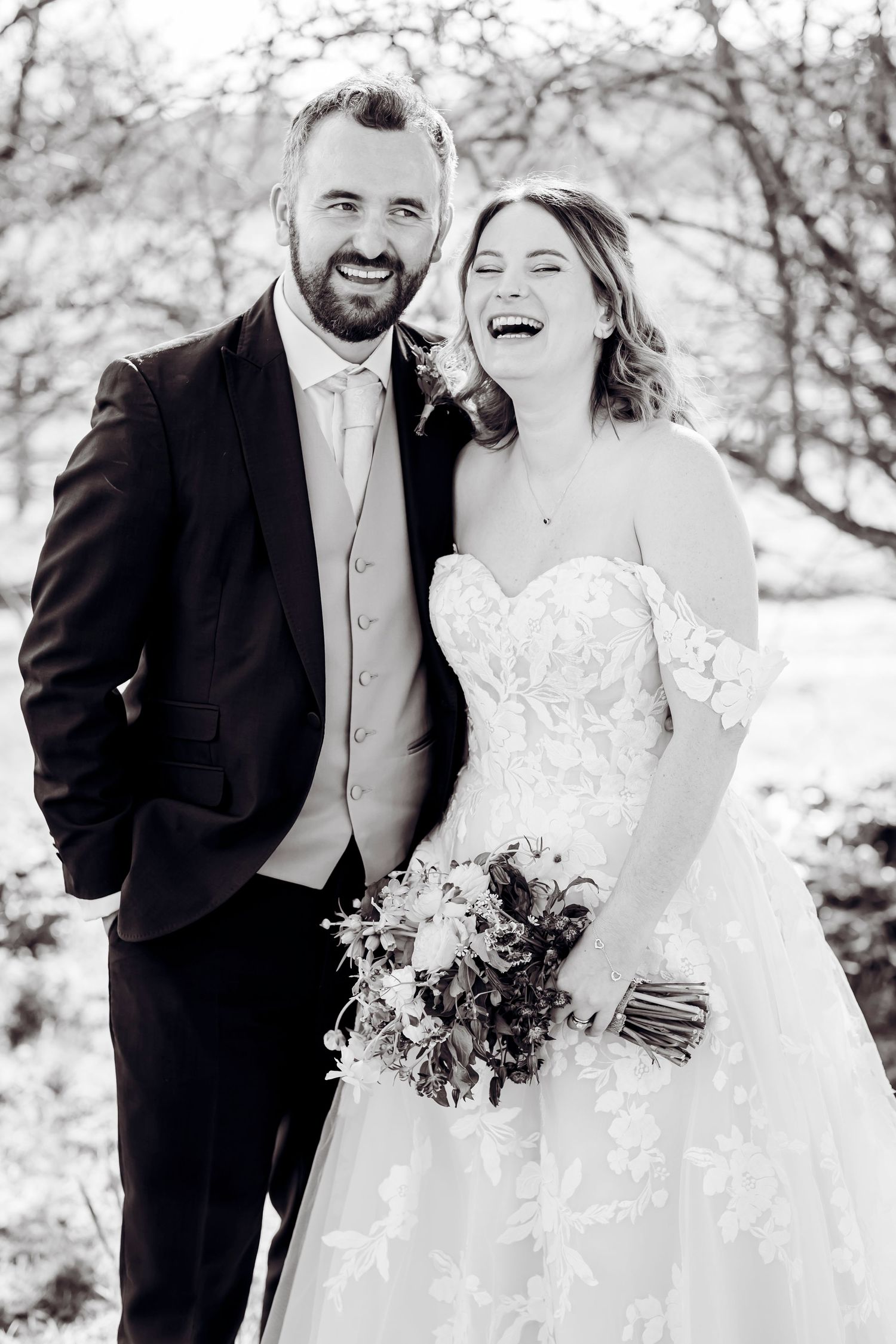 Black and white portrait of laughing bride and smiling groom holding bouquet outdoors.