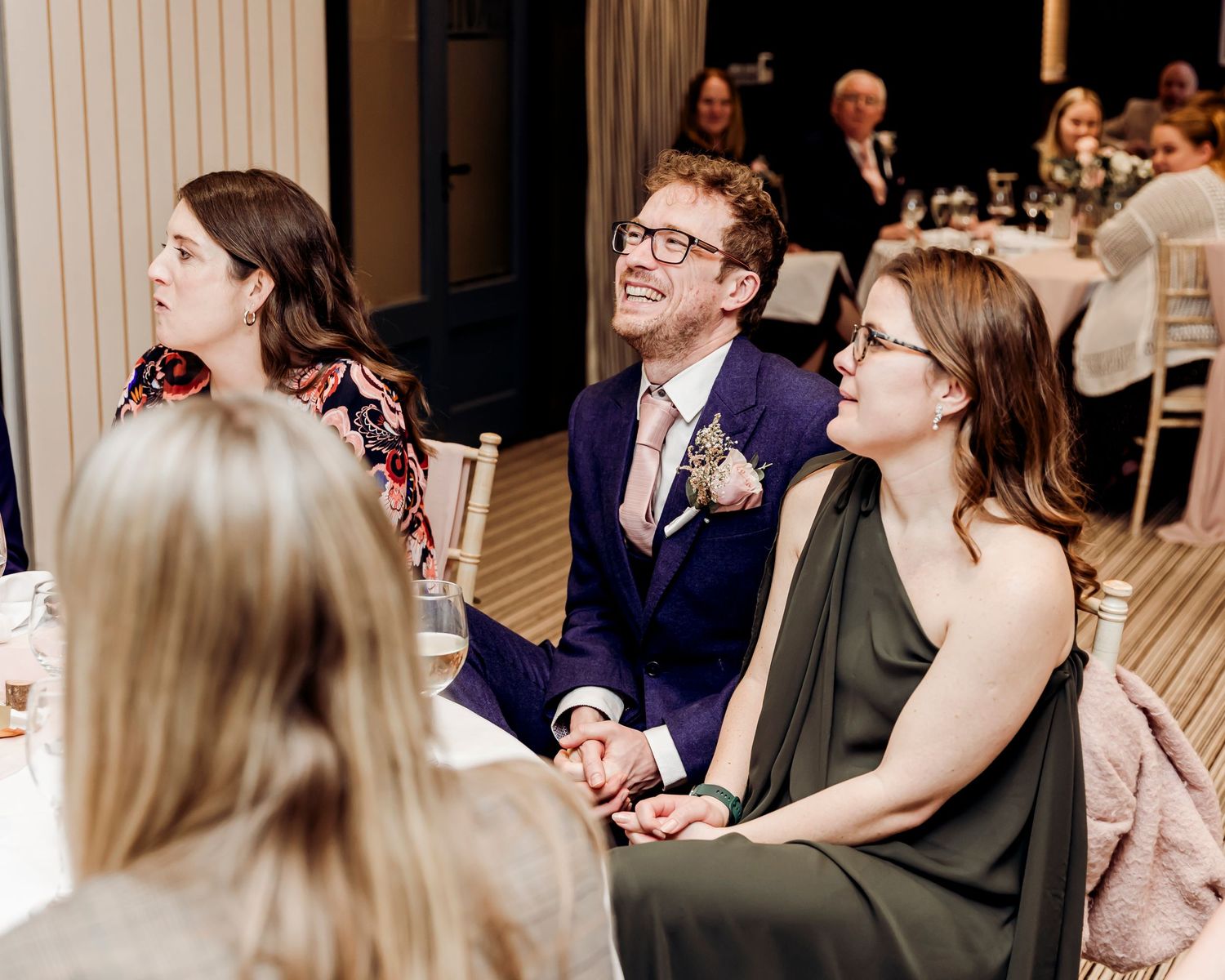 Wedding guests laughing and enjoying speeches at an elegant reception venue with fairy lights.