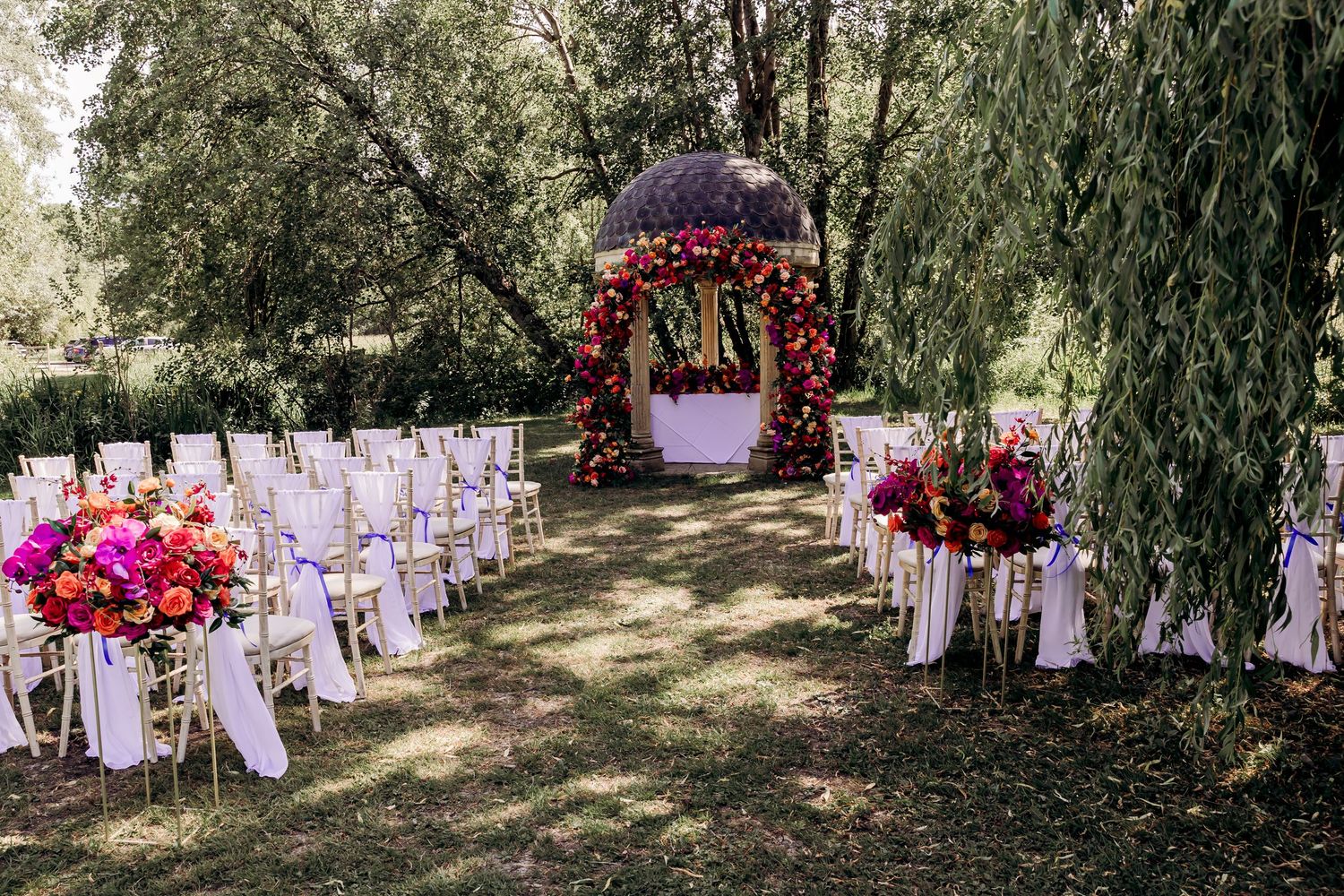Outdoor wedding ceremony setup with white chairs, vibrant floral arch, and colorful bouquets under lush green trees.